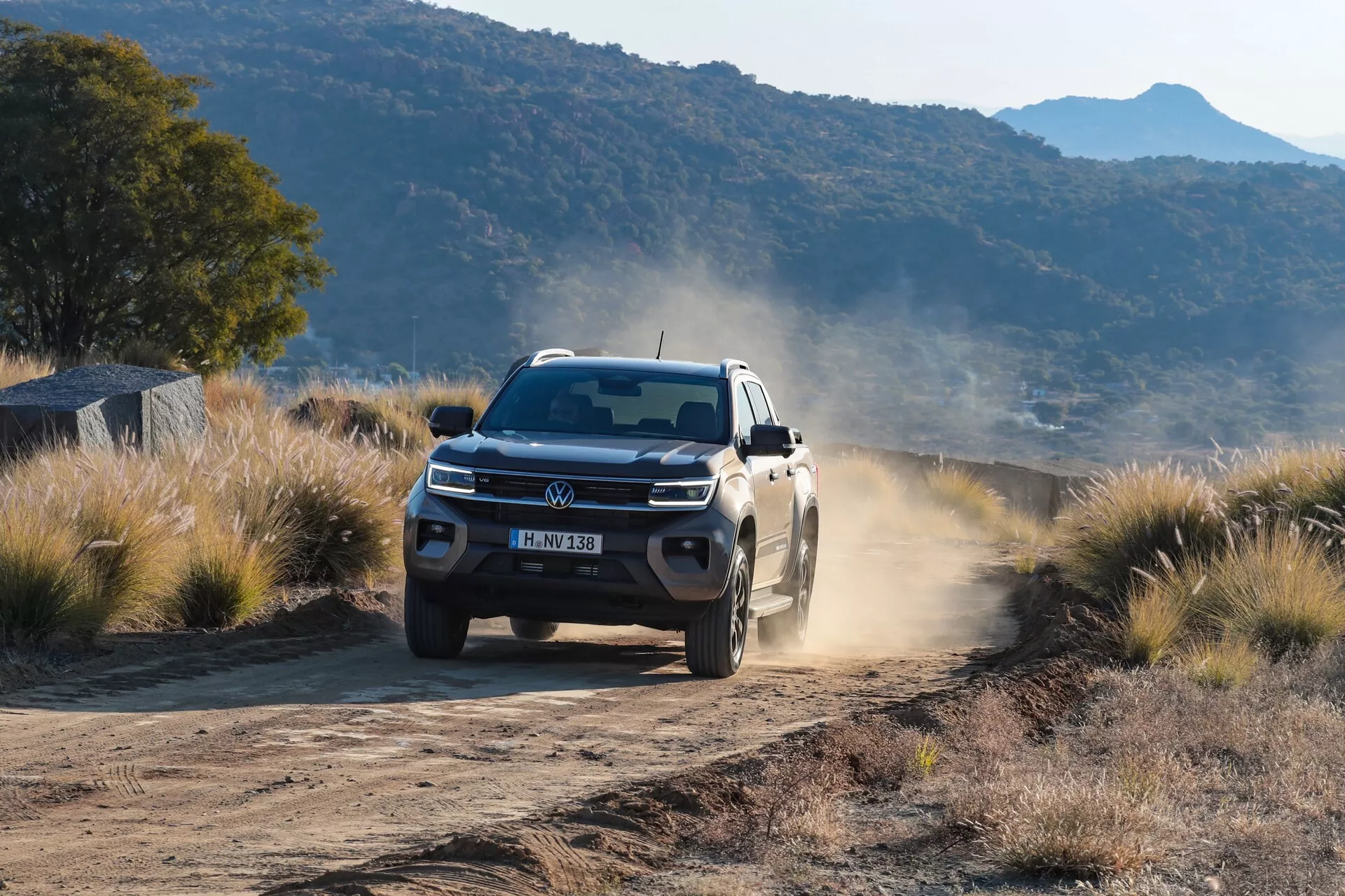 Un pick-up Volkswagen Amarok 2023 (2ème génération) blanc capturé en mouvement sur une route rurale non pavée et poussiéreuse, démontrant ses capacités tout-terrain. Le véhicule est montré sous un angle bas et avant, mettant en évidence le design robuste, les phares à LED, l'emblème VW, les jantes en alliage et la traînée poussiéreuse laissée derrière. La scène suggère tôt le matin ou tard dans la journée avec de plus longues ombres en dessous.