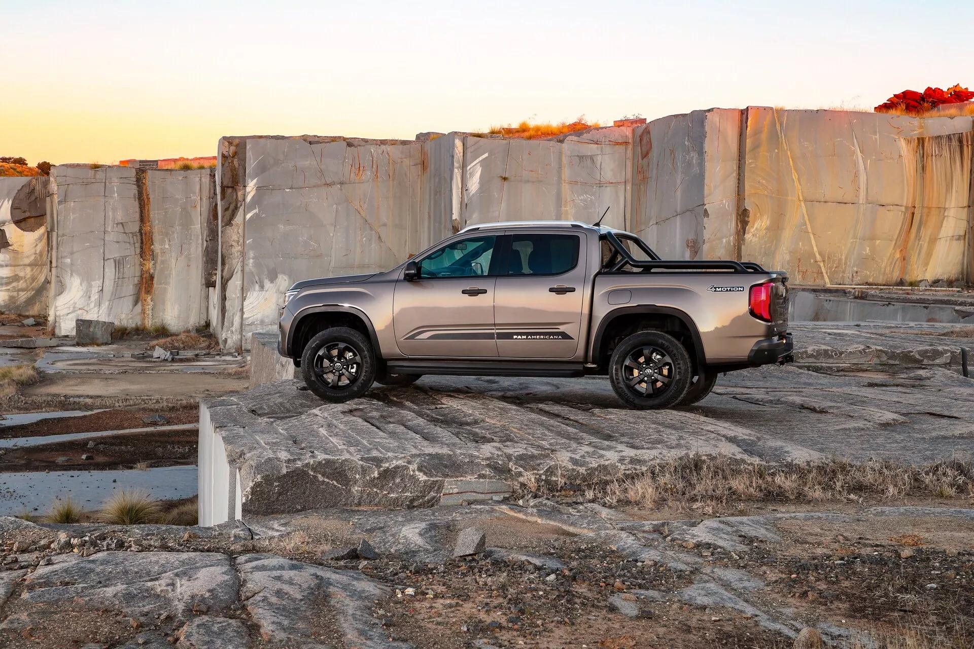Photographie d'un pick-up 2023 VW Nutzfahrzeuge Amarok dans un cadre extérieur, peut-être une carrière ou une zone avec de grandes dalles de pierre. L'image capture le camion à l'aube ou au crépuscule, mettant en valeur son carrosserie profilée, son design à double cabine, ses roues robustes tout-terrain, ses accents chromés et sa peinture métallisée. Le camion est positionné en diagonale avec l'avant légèrement incliné vers la caméra, montrant ses rétroviseurs extérieurs, ses caractéristiques ajustables et ses roues en alliage. L'environnement aride et la surface en pierre inégale soulignent la durabilité et les capacités tout-terrain du véhicule.