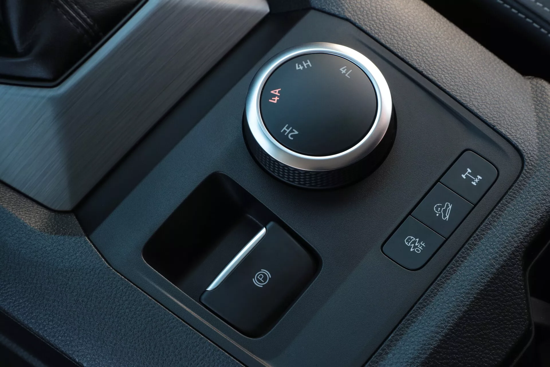 Vue rapprochée de l'intérieur du VW Nutzfahrzeuge Amarok 2023, mettant en avant le système de gestion du terrain et le sélecteur de mode de conduite, comprenant un cadran rotatif placé au centre avec des accents chromés indiquant des réglages de traction intégrale haute et basse. Les boutons à proximité incluent le contrôle de descente et la désactivation du contrôle de stabilité électronique, complétant une console au fini mat élégant avec un bouton d'alimentation sous le cadran pour l'allumage par bouton-poussoir.