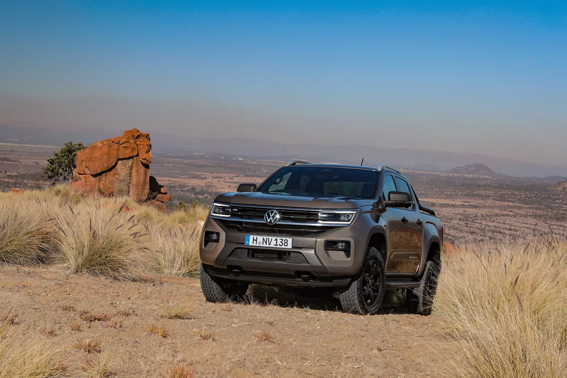 Un pick-up Volkswagen Amarok 2023 (2e génération) dans un environnement extérieur, tout-terrain, capturé sous un angle avant de trois quarts, montrant son côté passager avec une vue claire de la calandre avant, du pare-chocs, des phares, des feux antibrouillard, des passages de roue et une partie du profil latéral, y compris les portes passager et le rétroviseur latéral. La posture surélevée, la garde au sol élevée et la bande de roulement robuste soulignent ses capacités tout-terrain. Le cadre pittoresque du terrain accidenté et des collines lointaines met en valeur le design et le caractère aventureux du véhicule.