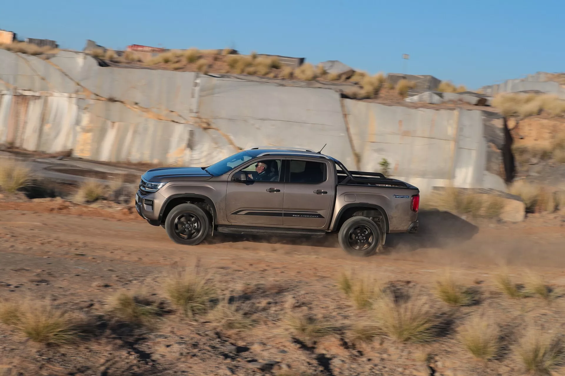 L'image montre une camionnette moyenne VW-Nutzfahrzeuge Amarok 2023 en mouvement, capturée sous un profil latéral dynamique dans des conditions d'éclairage chaudes en fin d'après-midi. Le camion roule sur une surface de terre, soulevant de la poussière des roues arrière, mettant en évidence ses capacités tout-terrain. Le véhicule présente des ailes prononcées, des moulures robustes autour des passages de roue, des marchepieds solides et des roues en alliage. Il a une configuration de cabine à quatre portes avec une caisse de chargement courte. L'arrière-plan flou, indiquant une carrière ou un site d'excavation, renforce l'effet dramatique.