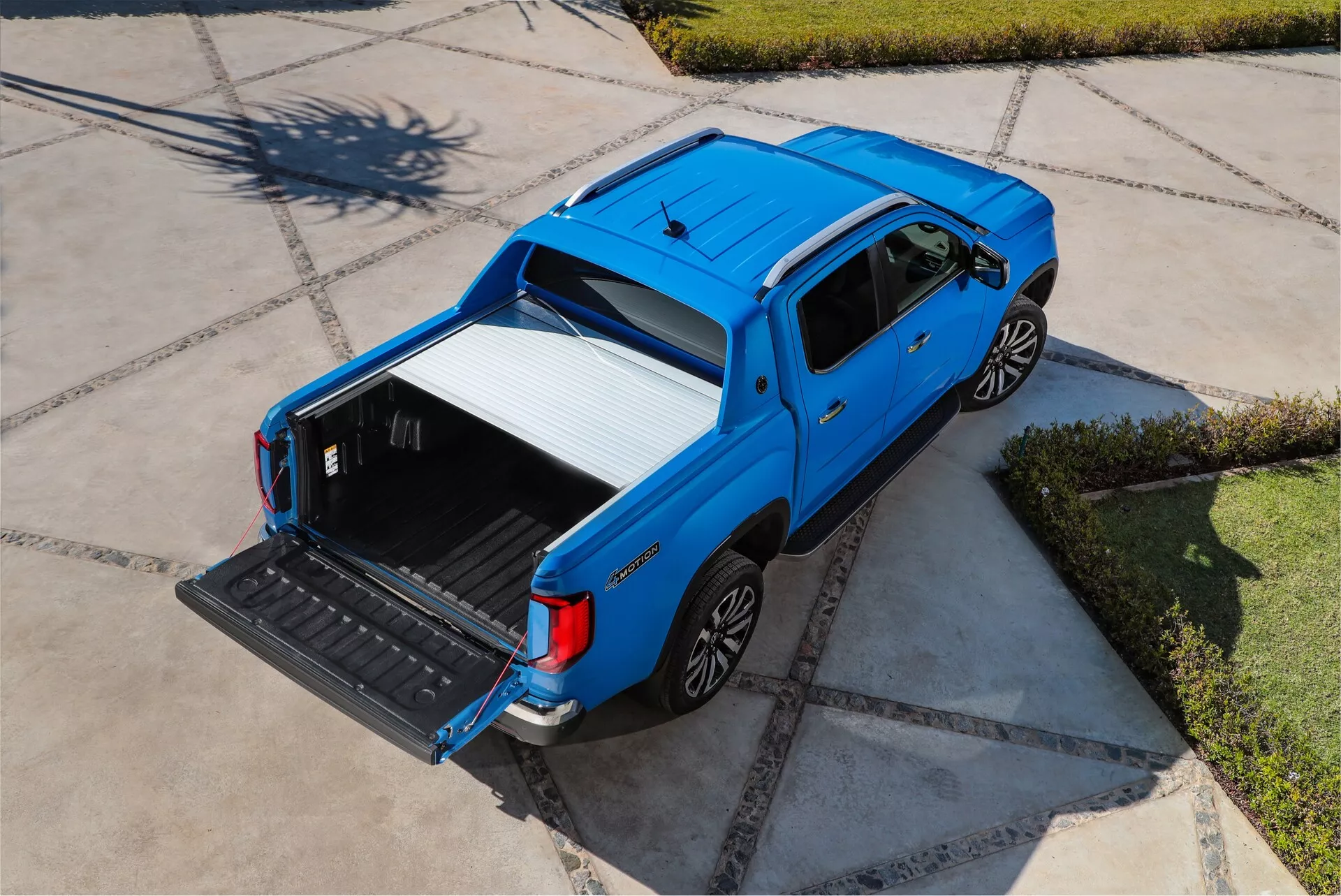 Image aérienne du pickup bleu VW-Nutzfahrzeuge Amarok 2e génération 2023, montrant son profil supérieur et latéral depuis un point de vue élevé. Le véhicule est stationné sur une allée pavée de manière distinctive, baignée de lumière solaire. Pris d'une perspective légèrement inclinée vers l'arrière, l'image met en évidence la benne ouverte avec la ridelle abaissée, révélant un revêtement protecteur durable. Le design élégant et contemporain à quatre portes est accentué par les rétroviseurs et les poignées de porte assortis à la couleur de la carrosserie. Les roues présentent un design en alliage multi-rayons, améliorant à la fois l'esthétique et les performances potentielles.