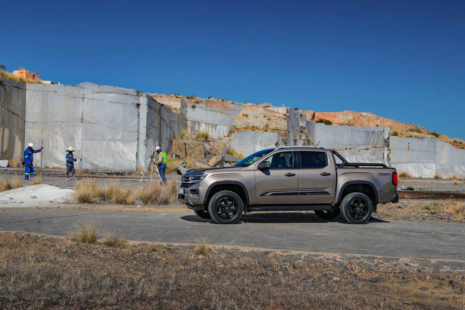 Vue de profil latéral du VW Nutzfahrzeuge Amarok 2023 de deuxième génération, garé sur une surface plane dans une carrière ou un site de construction, doté d'une cabine d'équipe avec des portes avant et arrière pleine grandeur, peinture mate grise ou beige, garnitures et roues noires, vitres teintées foncées, design moderne de la carrosserie avec élargisseurs d'ailes, marches latérales, possible barre de sport, phares LED, rétroviseurs grands, pneus tout-terrain et un vaste plateau de chargement avec couvre-tonneau. En arrière-plan, des personnes portant des équipements de sécurité.