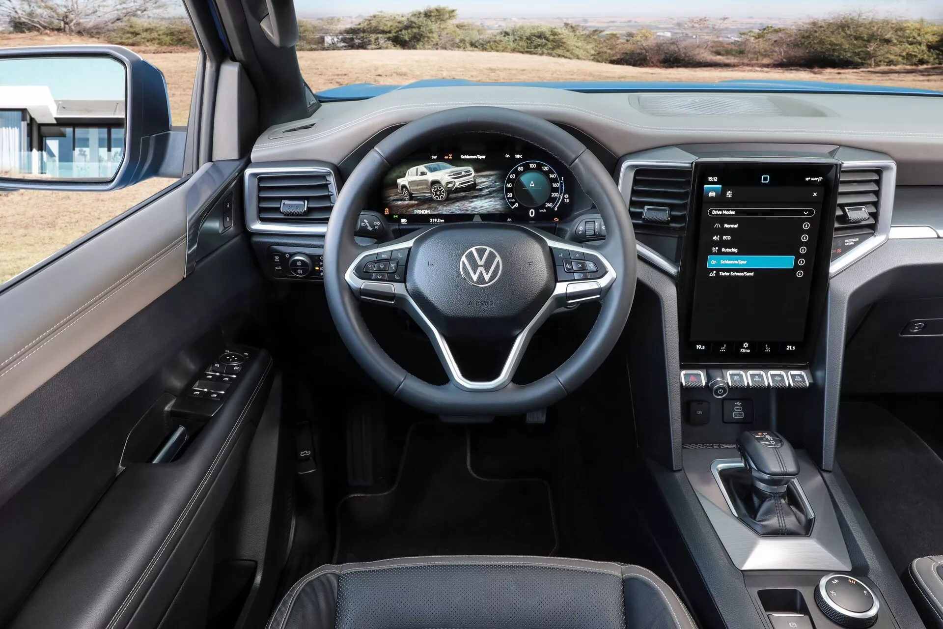 L'image montre l'intérieur d'un Volkswagen Nutzfahrzeuge Amarok 2023 (2e génération) depuis la perspective du passager avant. L'accent est mis sur le volant multifonctionnel arborant le logo Volkswagen. Derrière, se trouve un tableau de bord entièrement numérique indiquant la vitesse, les RPM et la navigation. La console centrale est dominée par un grand écran tactile vertical, qui offre les réglages du véhicule, les contrôles audio et l'intégration des smartphones comme Apple CarPlay ou Android Auto. En dessous, des commandes physiques minimales, un levier de vitesse élégant, un espace de rangement ouvert et un pad de recharge sans fil, indiquent un design moderne et avancé. L'intérieur présente une esthétique propre et contemporaine avec des surfaces douces au toucher, des accents en métal brossé et un haut-parleur central, ce qui suggère un système audio premium.