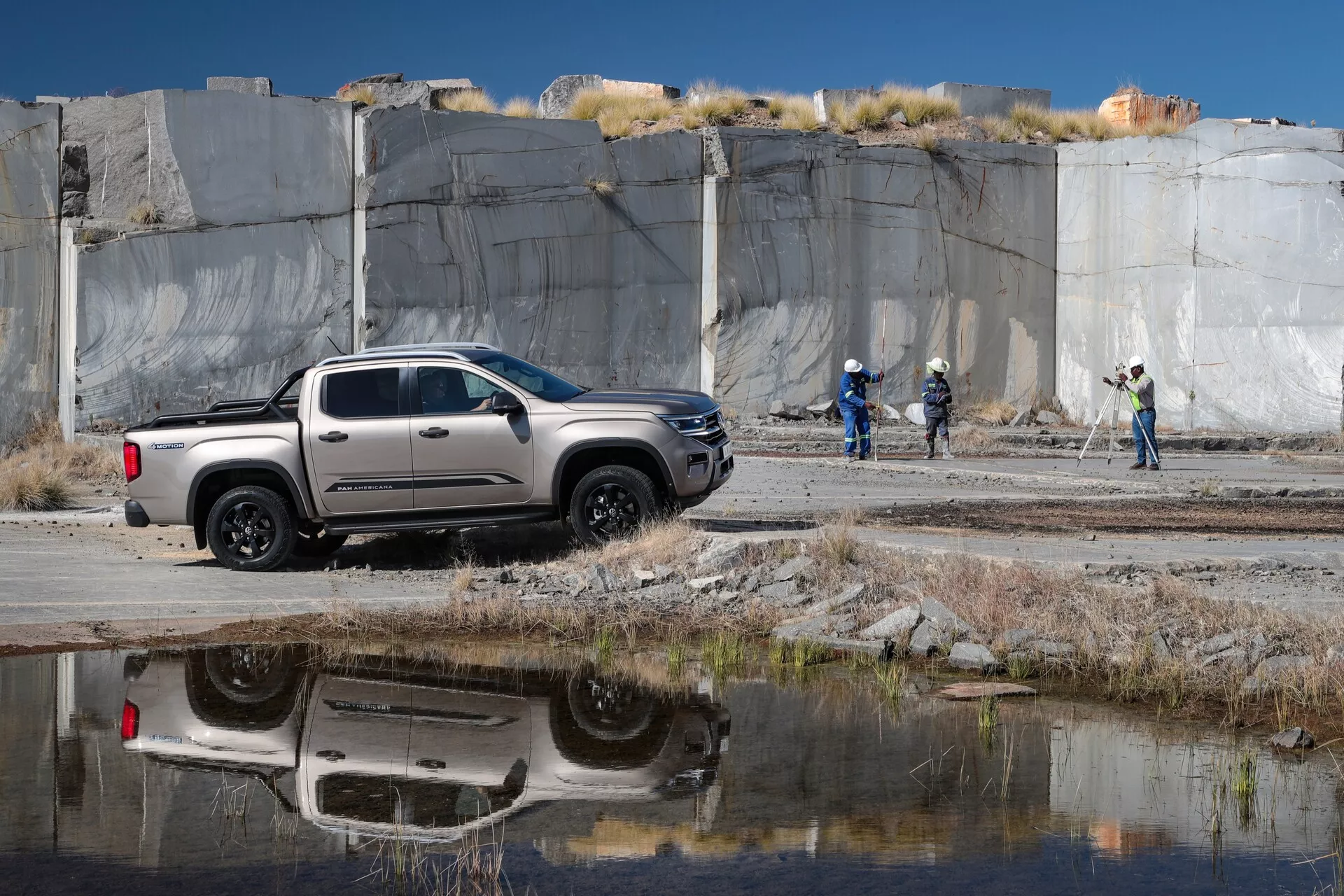 Un pick-up de taille moyenne VW Nutzfahrzeuge Amarok de 2023 en configuration crew cab est garé à un angle oblique avec l'avant face à droite. Capturé en profil latéral, le pick-up arbore clairement quatre portes, des rétroviseurs latéraux, des poignées de portes, des roues et des lignes de style sur une carrosserie de couleur neutre argentée ou gris clair avec des détails noirs autour des arches de roues et de la partie inférieure du corps. Le véhicule est sur une surface de gravier ou de terre avec des touffes d'herbe et une flaque d'eau reflétant son dessous. En arrière-plan se trouve une carrière ou un site minier avec des dalles de pierre et des travailleurs portant des casques et des gilets haute visibilité, suggérant un cadre industriel ou de construction. Les éléments technologiques notables incluent des roues en alliage et des rétroviseurs extérieurs, avec un design à haute garde au sol. L'image est prise en journée avec un éclairage naturel clair.