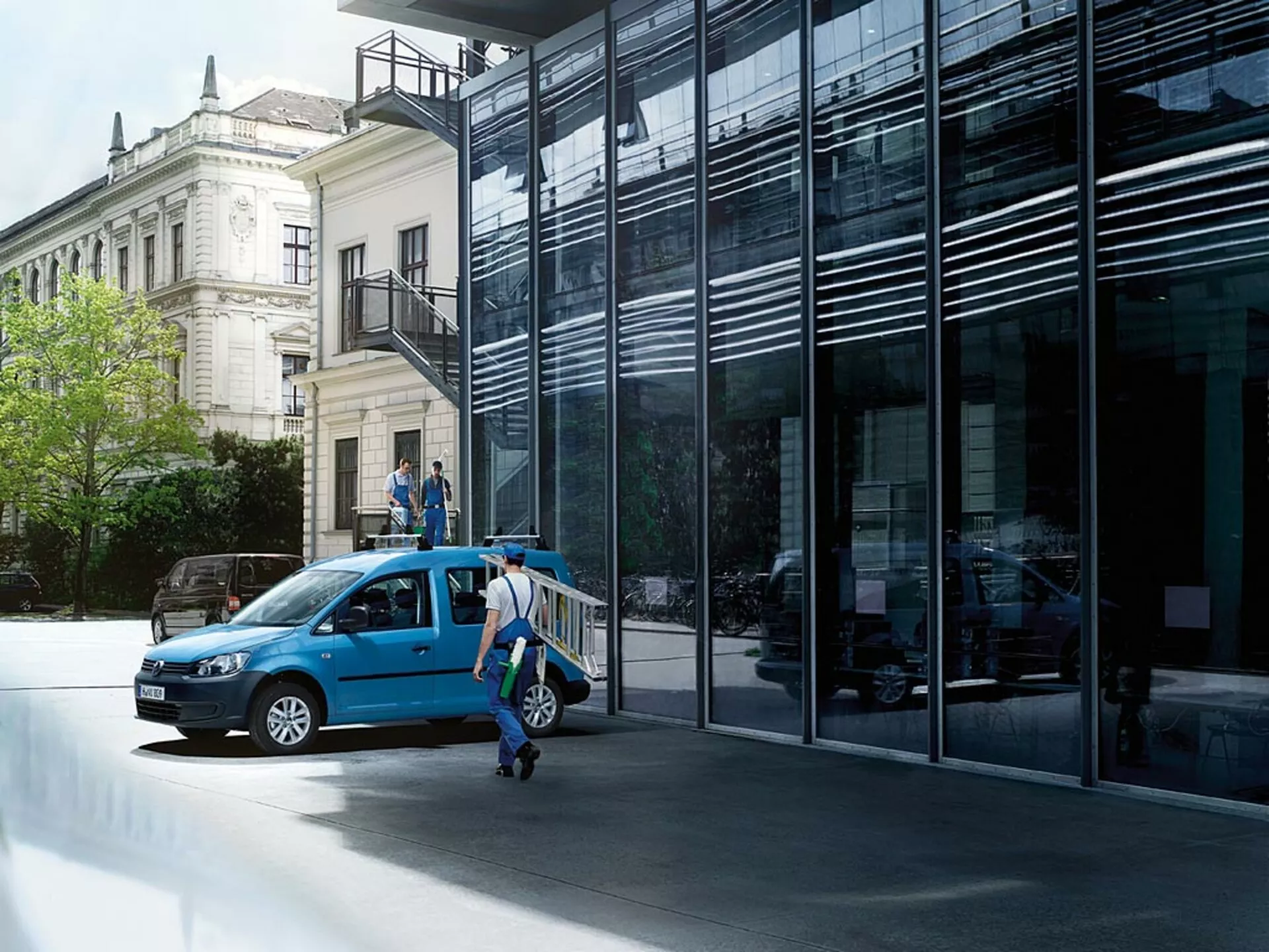 VW Caddy bleue de 2010 garée dans une rue urbaine, présentée dans une vue de trois quarts avant avec les portes ouvertes et mettant en exergue sa calandre avant, ses phares, ses roues, et un aperçu de l'intérieur sur fond d'architecture moderne et classique