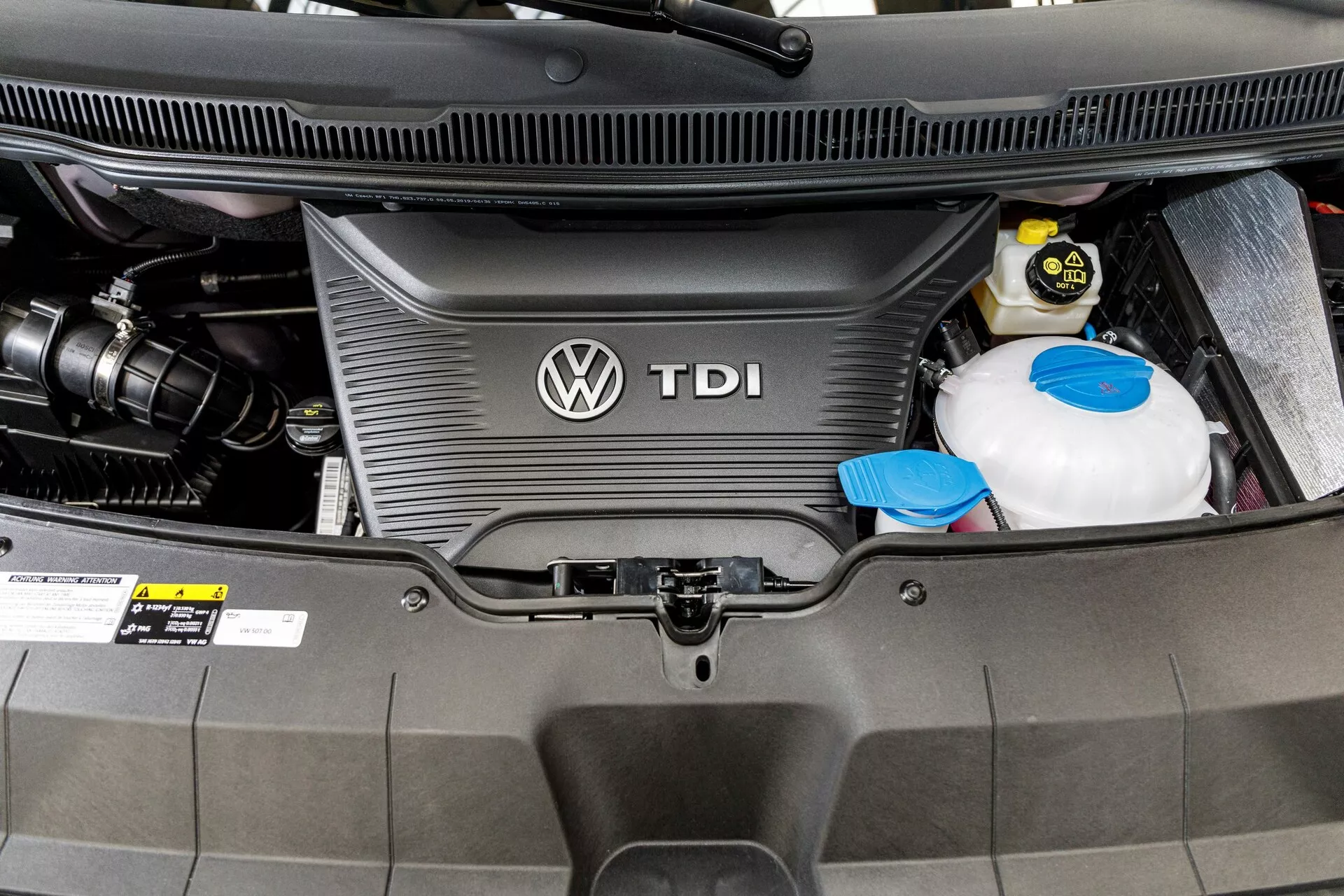 Vue rapprochée du compartiment moteur diesel du VW Nutzfahrzeuge Transporter 2019 (T6.1 California) montrant le couvercle du moteur TDI avec le logo Volkswagen, le réservoir de liquide de refroidissement et la batterie de la voiture