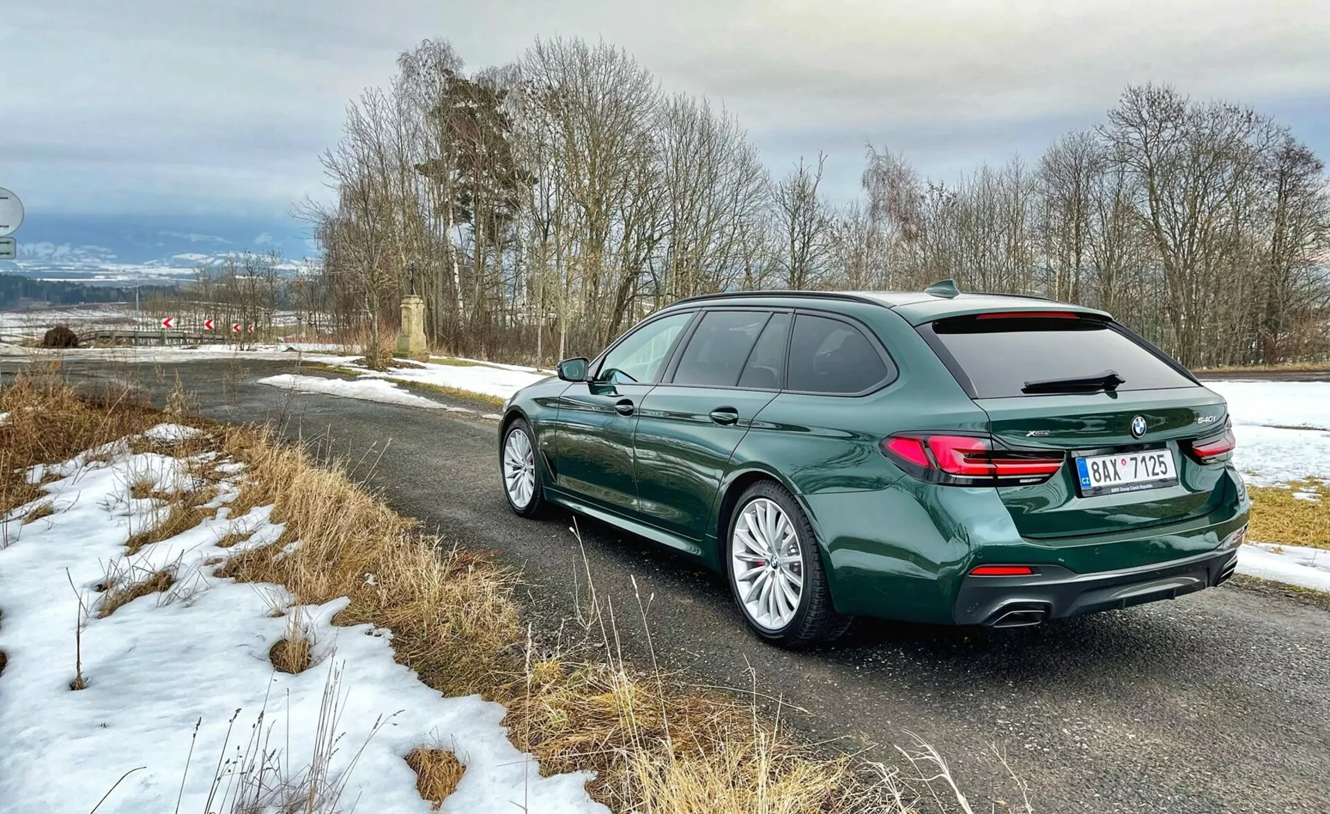 Vue arrière trois quarts d'un break BMW, montrant l'arrière du véhicule avec des feux arrière en forme de L, le profil latéral gauche comprenant les portes et la courbure du toit avec des barres de toit, jantes en alliage avec pneus routiers, en peinture verte foncée métallisée, garée sur une route de campagne avec un léger saupoudrage de neige, mettant en évidence des éléments de luxe et de design moderne.