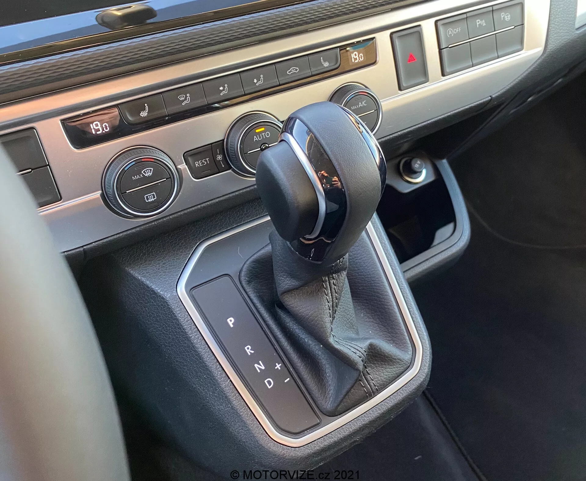 Vue rapprochée de la console centrale du VW Nutzfahrzeuge T6-1 Multivan 2019, centrée sur la zone du levier de vitesse. L'image montre un sélecteur de transmission automatique avec des coutures en cuir, entouré d'une garniture métallique, des boutons de contrôle de la climatisation avec affichage de la température et des commandes de chauffage des sièges, mettant en avant la technologie intérieure moderne et les matériaux haut de gamme du véhicule.