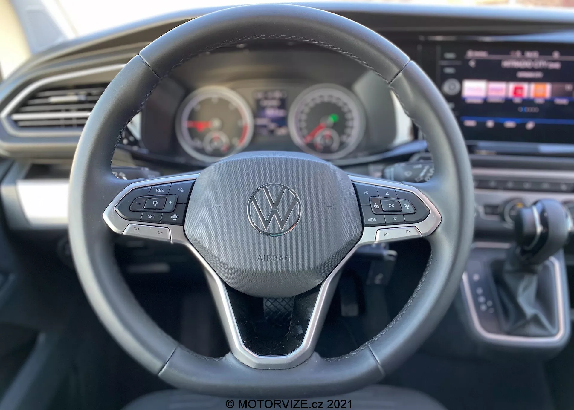 Vue intérieure d'un VW Nutzfahrzeuge Transporter 2019 (T6-1 Multivan) mettant l'accent sur la perspective du conducteur. Vue détaillée du volant avec le logo Volkswagen et des boutons de commande pour diverses fonctions telles que le régulateur de vitesse et la navigation multimédia. Derrière le volant, le tableau de bord présente un compteur de vitesse et un compte-tours de style analogique avec un affichage numérique au centre. Console centrale partiellement visible avec un grand écran tactile d'infodivertissement, accompagnée d'un design élégant du tableau de bord avec une grille d'aération horizontale. Levier de vitesses indiquant une transmission automatique à droite. L'image met en valeur les caractéristiques de convivialité avec une faible profondeur de champ se focalisant principalement sur le volant.