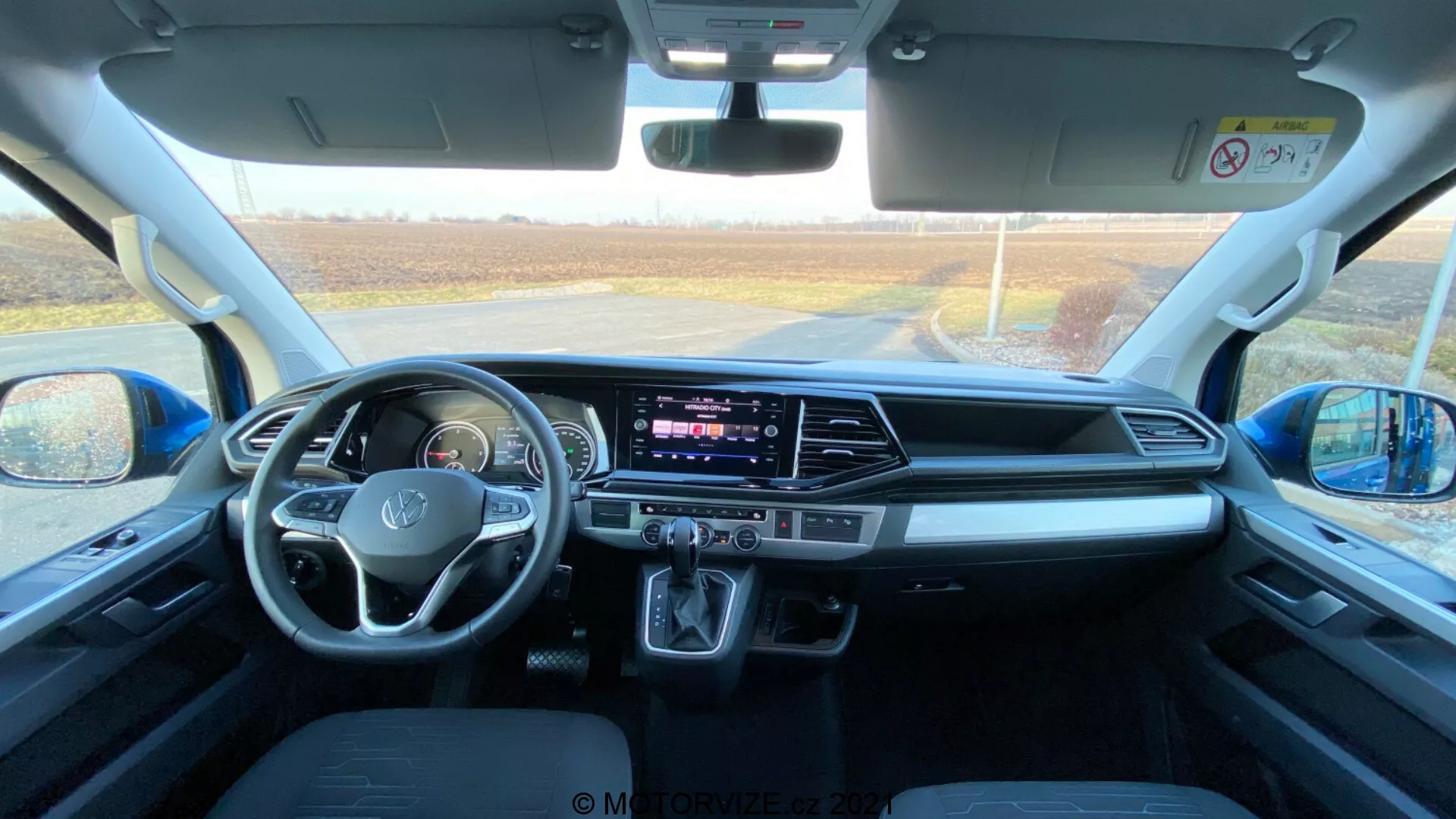 Intérieur d'un VW Nutzfahrzeuge Transporter 2019 (T6.1 Multivan), capturé depuis la zone des sièges arrière, regardant vers le tableau de bord avant. La photo est prise en journée avec la lumière naturelle entrant par le pare-brise avant, offrant une vue détaillée de l'espace de la cabine, des sièges avant, du volant avec commandes intégrées, de l'écran tactile central d'infodivertissement, des boutons et des boutons de la climatisation, du levier de vitesses automatique, des espaces de rangement, des ports de connectivité, des commandes de porte et de rétroviseur, des dispositifs de sécurité et du design confortable des sièges.