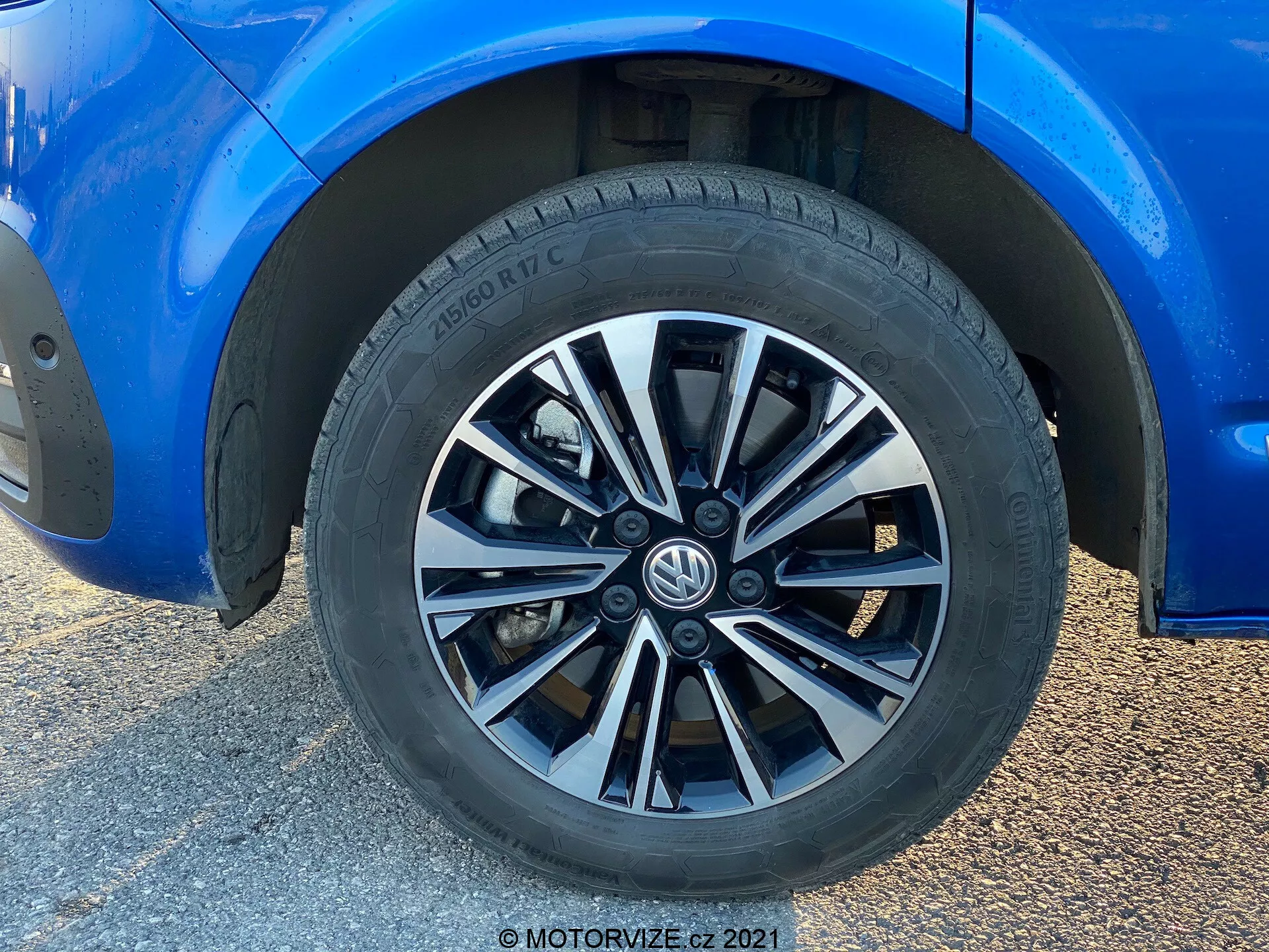 Gros plan d'une roue de Volkswagen T6-1 Multivan 2019. Ce cliché en plan rapproché présente une jante à multi-branches avec des finitions alternées argentées et foncées. Le pneu, de marque Hankook, révèle un motif de bande de roulement pour la performance sur route et des spécifications partielles. La carrosserie bleue environnante est en excellent état, montrant une brillance réfléchissante. Une garde au sol modérée et des étriers de frein standard partiellement visibles sont indiqués. La lumière naturelle met en valeur la composition centrée sur la roue, l'arrière-plan étant flou en raison de la prise de vue en contre-plongée.