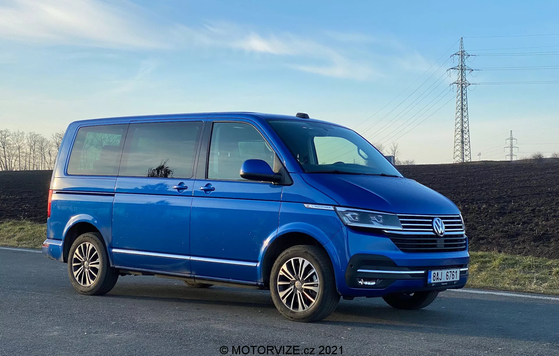 Une camionnette Volkswagen Transporter T6 bleue, montrant son profil latéral d'un angle bas, mettant en valeur sa longueur et sa hauteur. L'image présente des détails tels que la porte coulissante passager, la porte avant passager, la roue arrière côté passager, des jantes en alliage multi-branches et des vues partielles du phare, de la calandre Volkswagen avec emblème et du pare-chocs avant. La photo, prise en fin d'après-midi, montre la garde au sol et reflète le ciel et les environs dans les fenêtres, avec un ciel dégagé et des lignes électriques en arrière-plan.