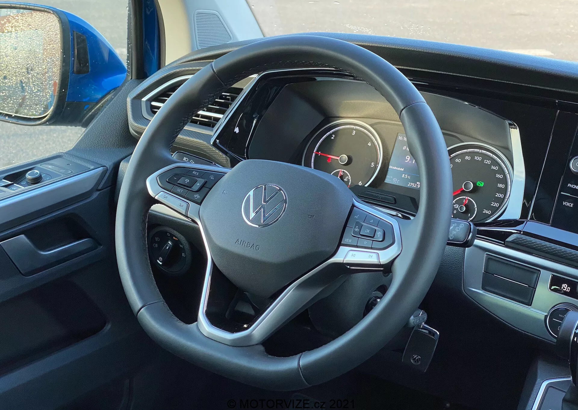 Vue intérieure du VW Nutzfahrzeuge Transporter 2019 (T6-1 Multivan) depuis la perspective du conducteur, centrée sur le volant et le tableau de bord. Le volant arbore l'emblème Volkswagen et est doté d'une finition en cuir, de boutons multifonctions et de palettes de changement de vitesse. Le combiné d'instruments partiellement visible comprend des cadrans analogiques et un écran numérique. L'écran central d'infodivertissement et les commandes de climatisation sont partiellement visibles, indiquant des fonctionnalités modernes de multimédia et de confort.