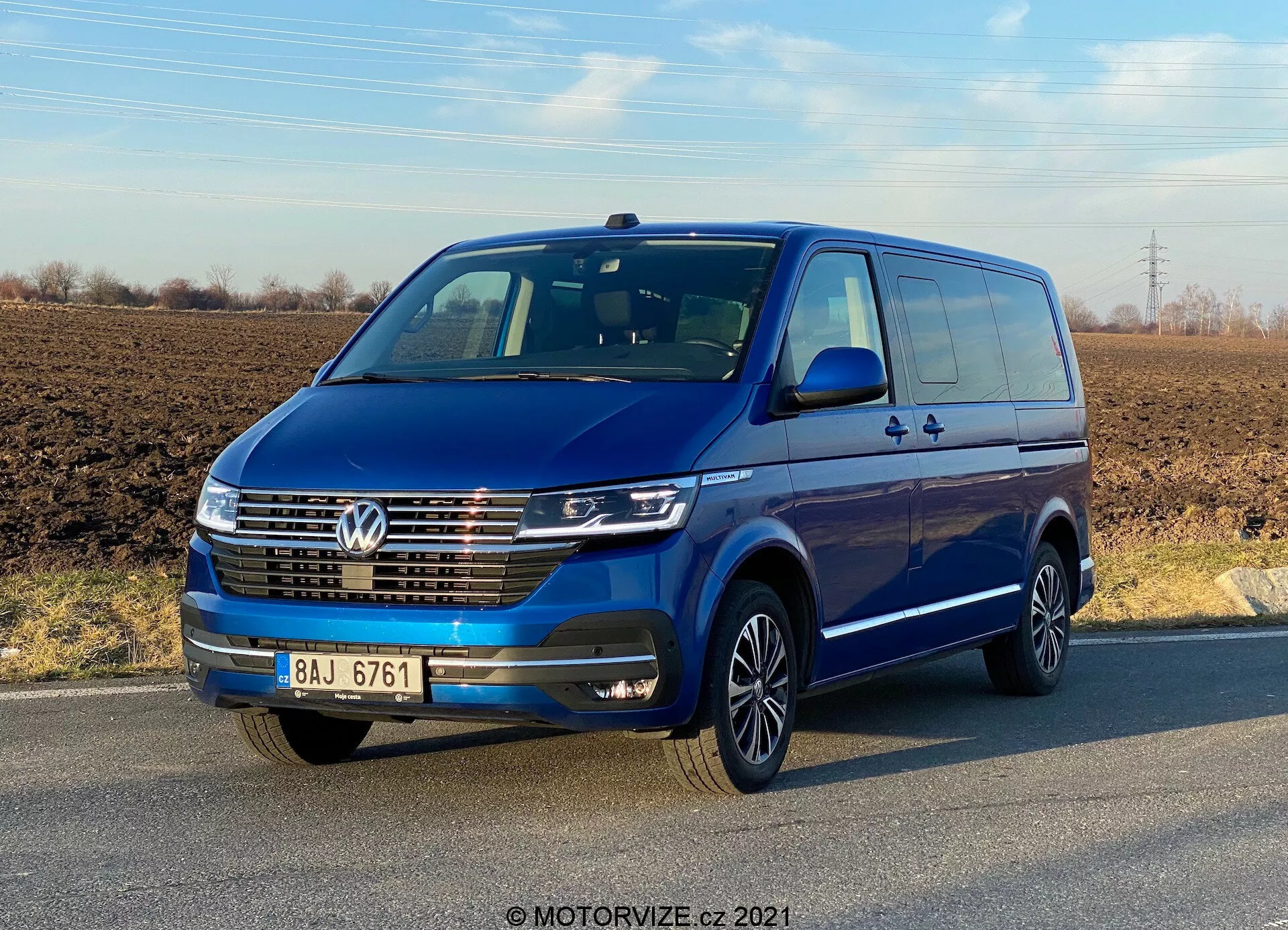 Vue de trois quarts avant d'un Volkswagen Transporter T6.1 Multivan bleu de 2019 sous un ciel dégagé. Le véhicule présente un logo Volkswagen sur la calandre, des feux de jour à LED, une garniture chromée sur la calandre avant et des jantes en alliage à plusieurs rayons. Les rétroviseurs et les pare-chocs de la couleur de la carrosserie suggèrent un modèle haut de gamme. Les portes et les poignées, y compris une porte coulissante arrière, sont visibles, indiquant sa capacité pour plusieurs passagers ou du fret.