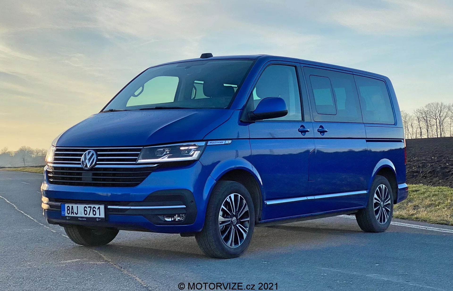 Image d'une fourgonnette bleue Volkswagen Transporter T6.1 Multivan 2019, prise sous un angle bas pour accentuer sa stature et sa taille. La vue de trois quarts avant montre le design frontal et latéral du véhicule, y compris la calandre distinctive Volkswagen avec des lignes horizontales, l'emblème central, les phares modernes probablement équipés de technologies LED ou Xenon, et un pare-chocs avant élégant avec une large prise d'air. Les rétroviseurs latéraux aérodynamiques avec clignotants intégrés, l'espace vitré généreux avec une porte latérale coulissante, les jantes en alliage avec des pneus à profil bas et un éclairage doux mettent en valeur la praticité et l'attrait esthétique de la fourgonnette. Le filigrane 'MOTORVIZE.cz 2021' suggère que l'image date de 2021.