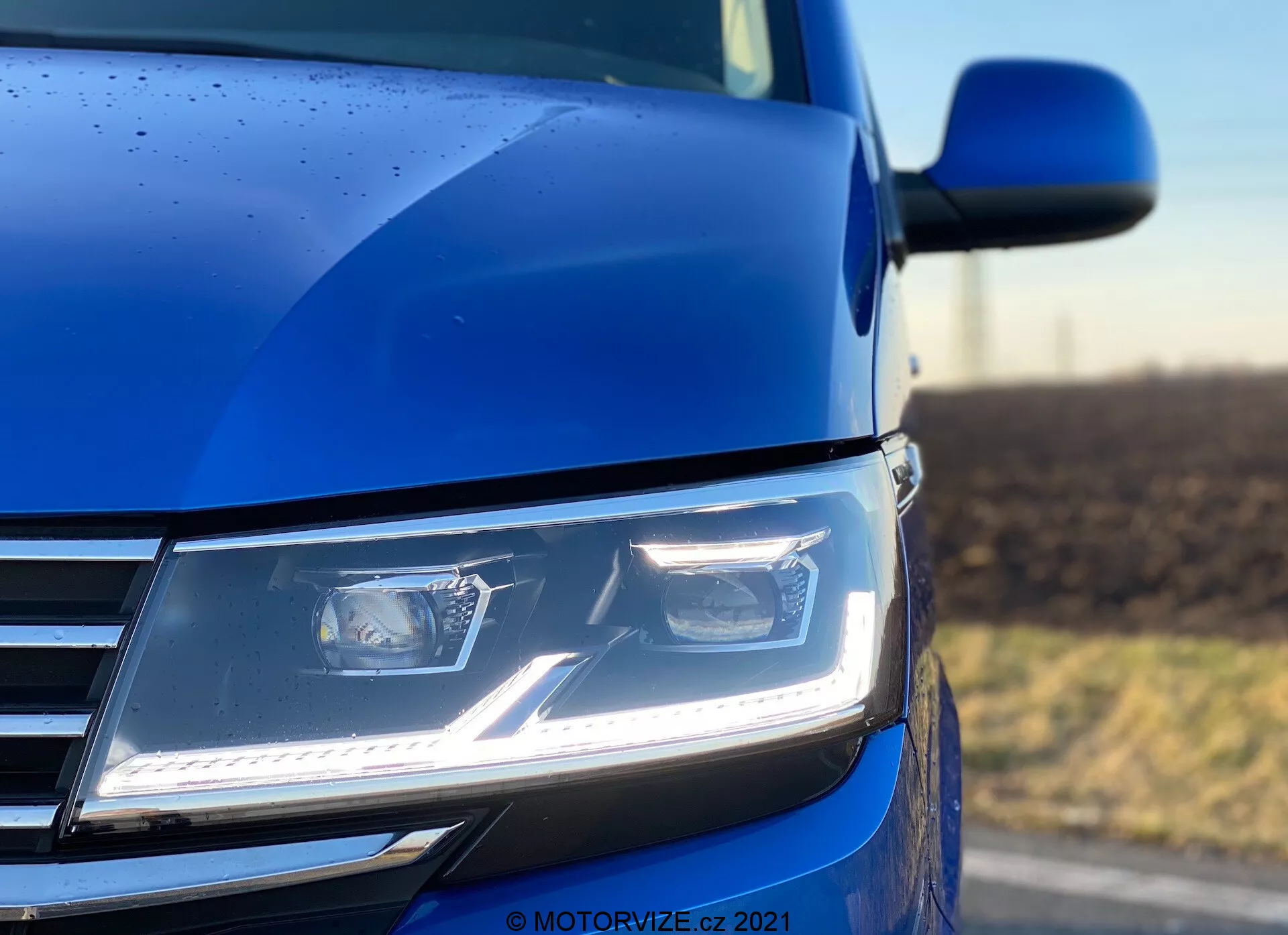Une vue rapprochée en angle bas de la section avant gauche du VW Nutzfahrzeuge Transporter 2019 (T6.1 Multivan) bleu, mettant en évidence les phares modernes à LED angulaires avec feux diurnes, une partie de la calandre chromée et un aperçu du contour du capot et du rétroviseur latéral. La photo, prise avec une faible profondeur de champ, met l'accent sur le phare et la carrosserie environnante, soulignant la technologie d'éclairage avancée, le design contemporain et la qualité de la peinture, complétée par des gouttelettes de pluie indiquant une finition extérieure détaillée.