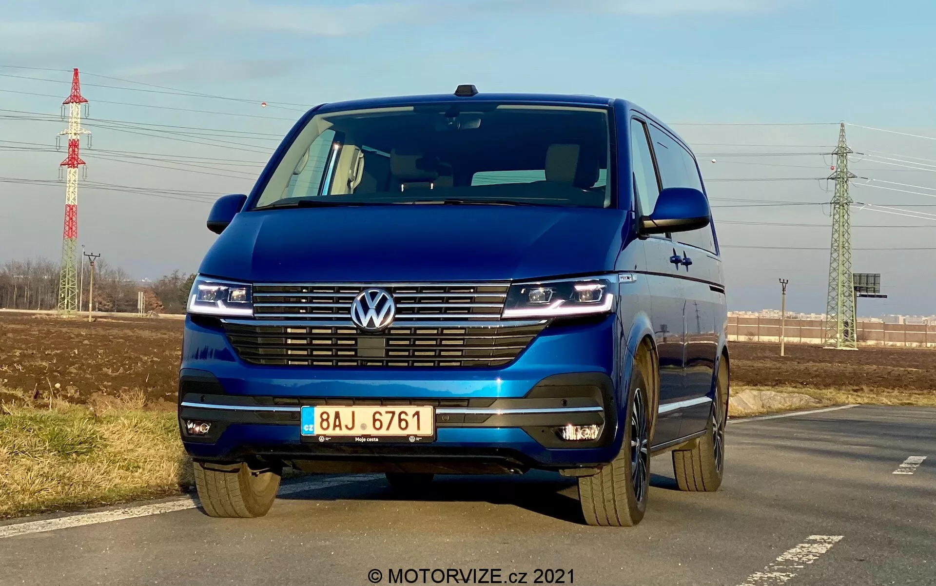 Volkswagen T6-1 Multivan 2019 bleu capturé sous un angle frontal bas mettant en avant la grille, les phares LED avec feux de jour, l'emblème Volkswagen, la large calandre, le pare-chocs bi-ton et les jantes en alliage; garé au bord d'une route avec un ciel clair, une verdure lointaine, un pylône électrique et des infrastructures en arrière-plan.