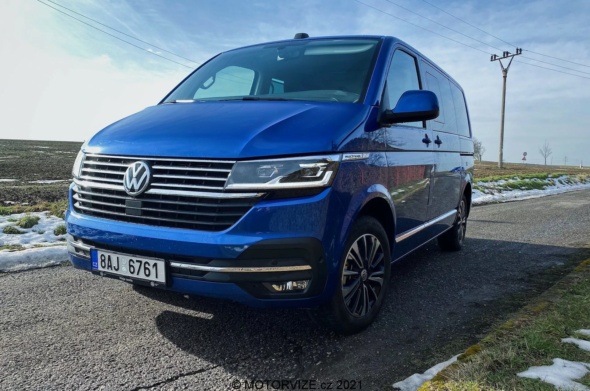 Volkswagen T6.1 Multivan Transporter 2019 bleu capturé en angle avant trois-quarts bas, montrant la face avant de la voiture, grille avec logo Volkswagen proéminent, barres horizontales chromées, phares LED angulaires avec feux de jour, pare-chocs avant avec antibrouillards intégrés et entrées d'air, ligne dynamique s'étendant sur les côtés, roues en alliage à plusieurs branches, clignotants intégrés aux rétroviseurs latéraux, vitres teintées. Le véhicule est garé sur une route mouillée à côté d'herbe enneigée, avec un arrière-plan rural comprenant des poteaux électriques et un ciel ouvert.