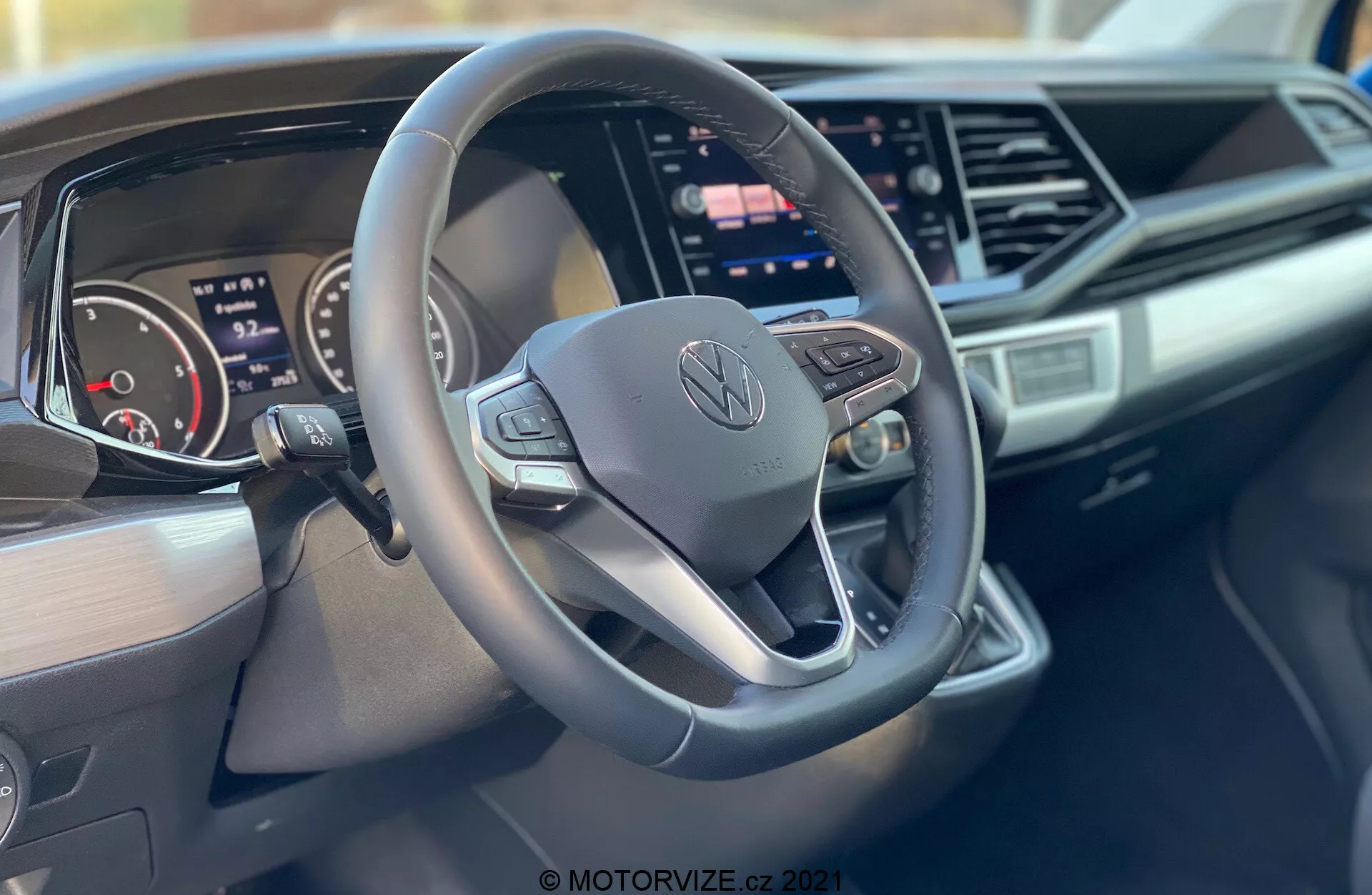 Vue intérieure d'un Volkswagen Nutzfahrzeuge Transporter 2019 (T6-1 Multivan) se concentrant sur le volant et le tableau de bord. Le gros plan pris du côté passager met en évidence un volant à trois branches au bas plat avec des boutons de commande et le logo central de Volkswagen, un tableau de bord numérique, un grand écran tactile d'info-divertissement, un design de tableau de bord élégant avec des accents angulaires et le branding côté passager du tableau de bord.