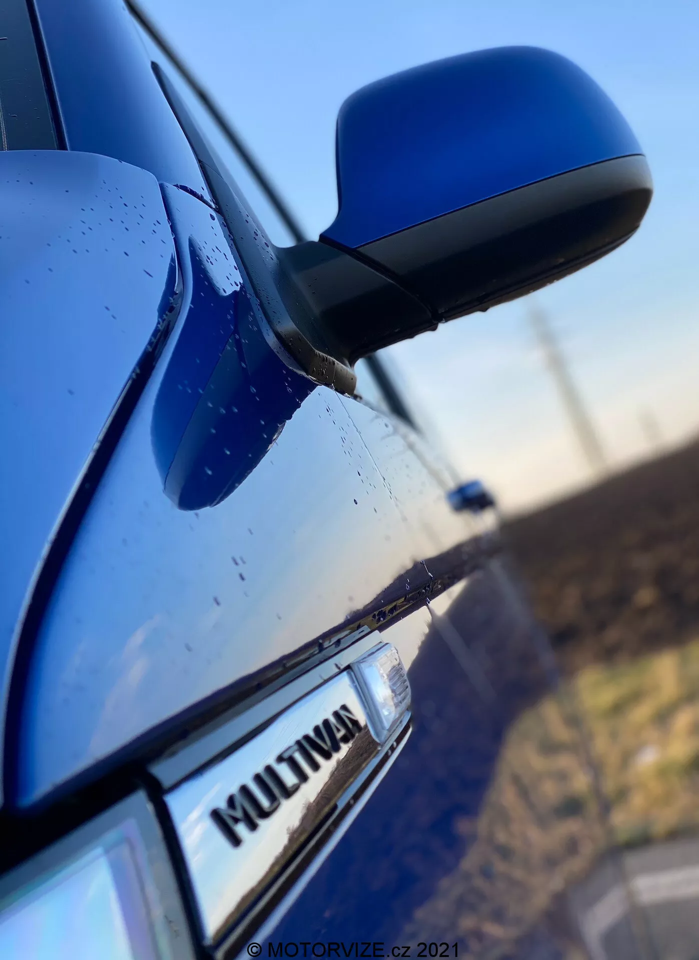 Gros plan du côté d'un VW Nutzfahrzeuge Transporter 2019 bleu (T6-1-Multivan) comprenant une partie de la porte passager avant, le rétroviseur latéral et une portion de la porte arrière. La mise au point est sur l'insigne 'MULTIVAN' de la voiture, avec le rétroviseur latéral aérodynamique légèrement flou. Le boîtier du rétroviseur est assorti à la couleur de la carrosserie, et des gouttelettes d'eau sont visibles sur la surface, suggérant des caractéristiques d'étanchéité ou une pluie récente.