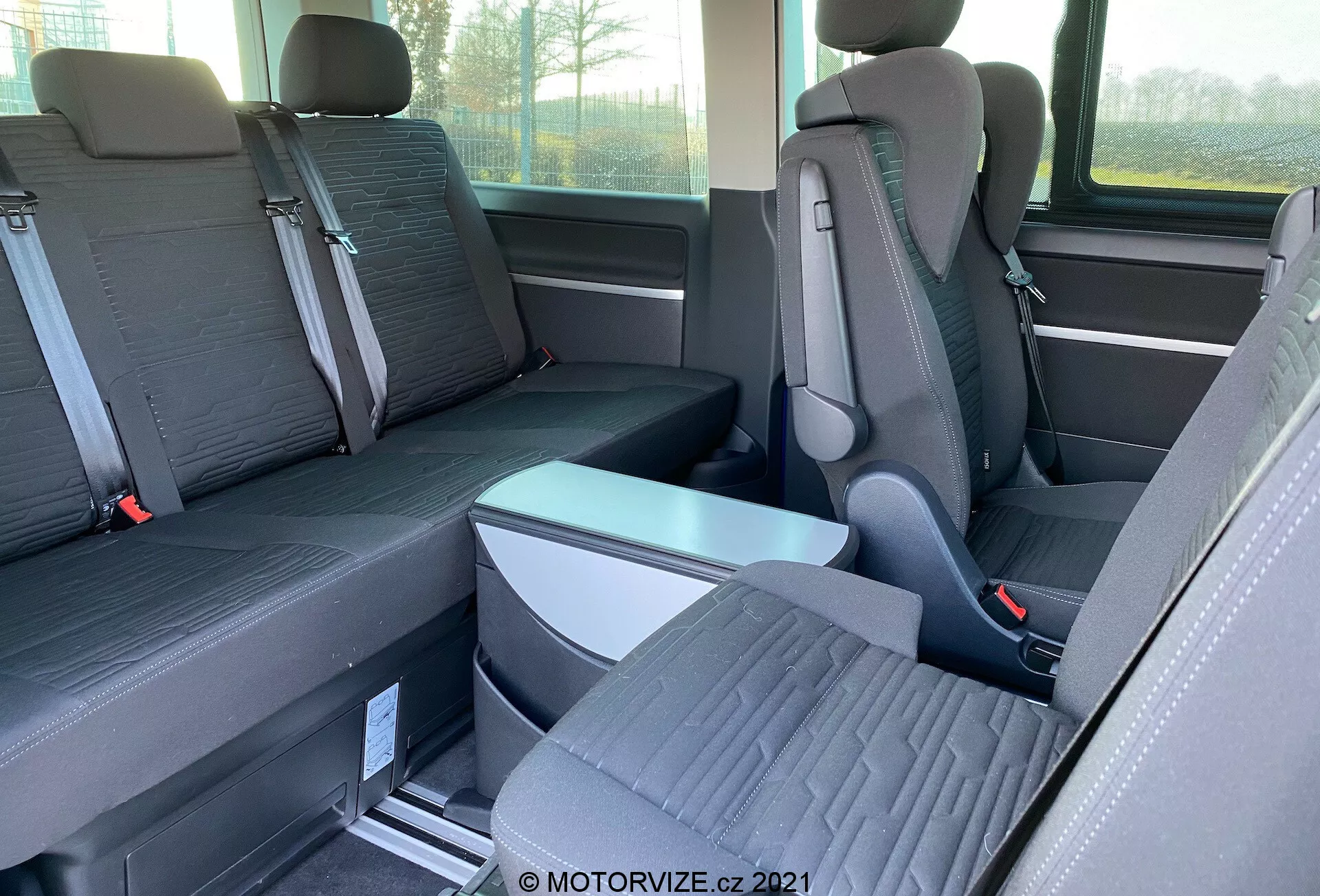 Vue intérieure du VW-Nutzfahrzeuge Transporter 2019 (T6.1 Multivan) se concentrant sur les sièges des deuxième et troisième rangées. Photographie prise de côté à hauteur d'œil, l'image montre des sièges individuels avec appuie-tête proéminents dans la deuxième rangée et une banquette pour trois passagers dans la troisième rangée. Les sièges sont en tissu bicolore et le plancher présente des rails métalliques pour des sièges reconfigurables. Un accoudoir pliable et des boucles de ceinture de sécurité sont visibles. La fenêtre et le rangement latéral sont également partiellement visibles.