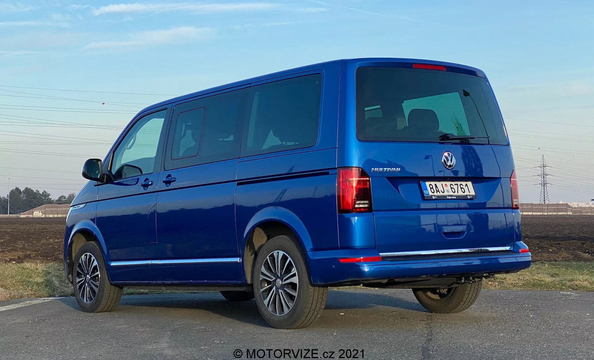 Une fourgonnette bleue VW-Nutzfahrzeuge Transporter 2019 (T6-1 Multivan) vue sous un angle arrière de trois quarts. L'image capture les groupes de feux arrière, les vitres arrière et le hayon ainsi que le profil latéral avec les portes et les fenêtres des passagers. La fourgonnette occupe une grande partie du cadre à hauteur des yeux, révélant des portes arrière divisées et plusieurs fenêtres latérales, indiquant un modèle pour passagers. Les roues en alliage, les pare-chocs couleur carrosserie et les rétroviseurs latéraux suggèrent un niveau de finition élevé. L'éclairage suggère une journée ensoleillée avec un ciel dégagé et des ombres nettes sous le véhicule.