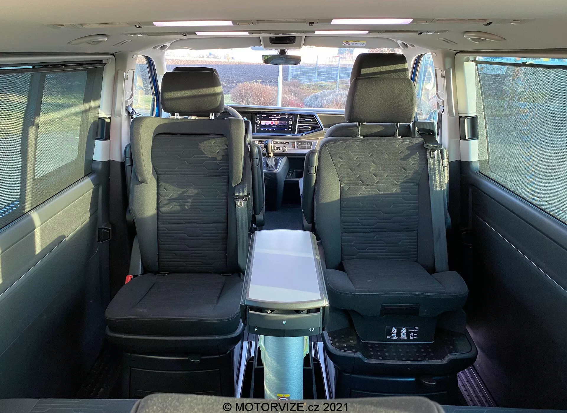 Intérieur du VW Nutzfahrzeuge Transporter T6-1 Multivan 2019 vu de l'arrière, montrant des sièges en cuir, des accoudoirs à la deuxième rangée, une table pliante, un toit panoramique, des sorties d'air individuelles et un système d'infodivertissement avancé ; met en avant le confort des passagers et l'habitacle spacieux.