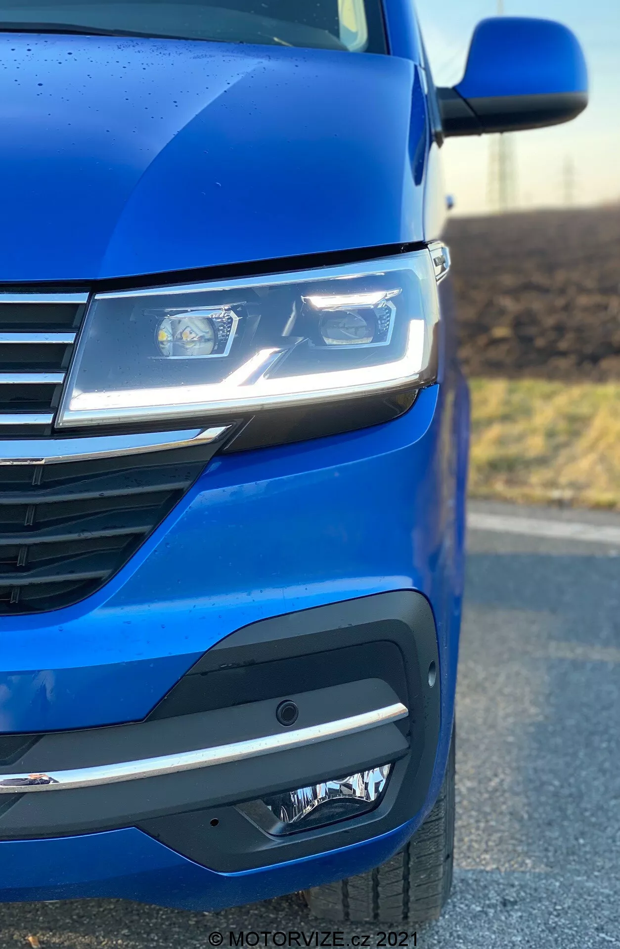 Gros plan sur le côté avant gauche d'un VW Nutzfahrzeuge Transporter 2019 (T6.1 Multivan) soulignant la peinture bleu métallique, les phares LED avancés, le design moderne de la grille avec des lignes horizontales et le pare-chocs avant sophistiqué avec des phares antibrouillard ou des unités de capteur. L'angle de la photo met en évidence l'aérodynamisme et les détails de conception de la voiture.