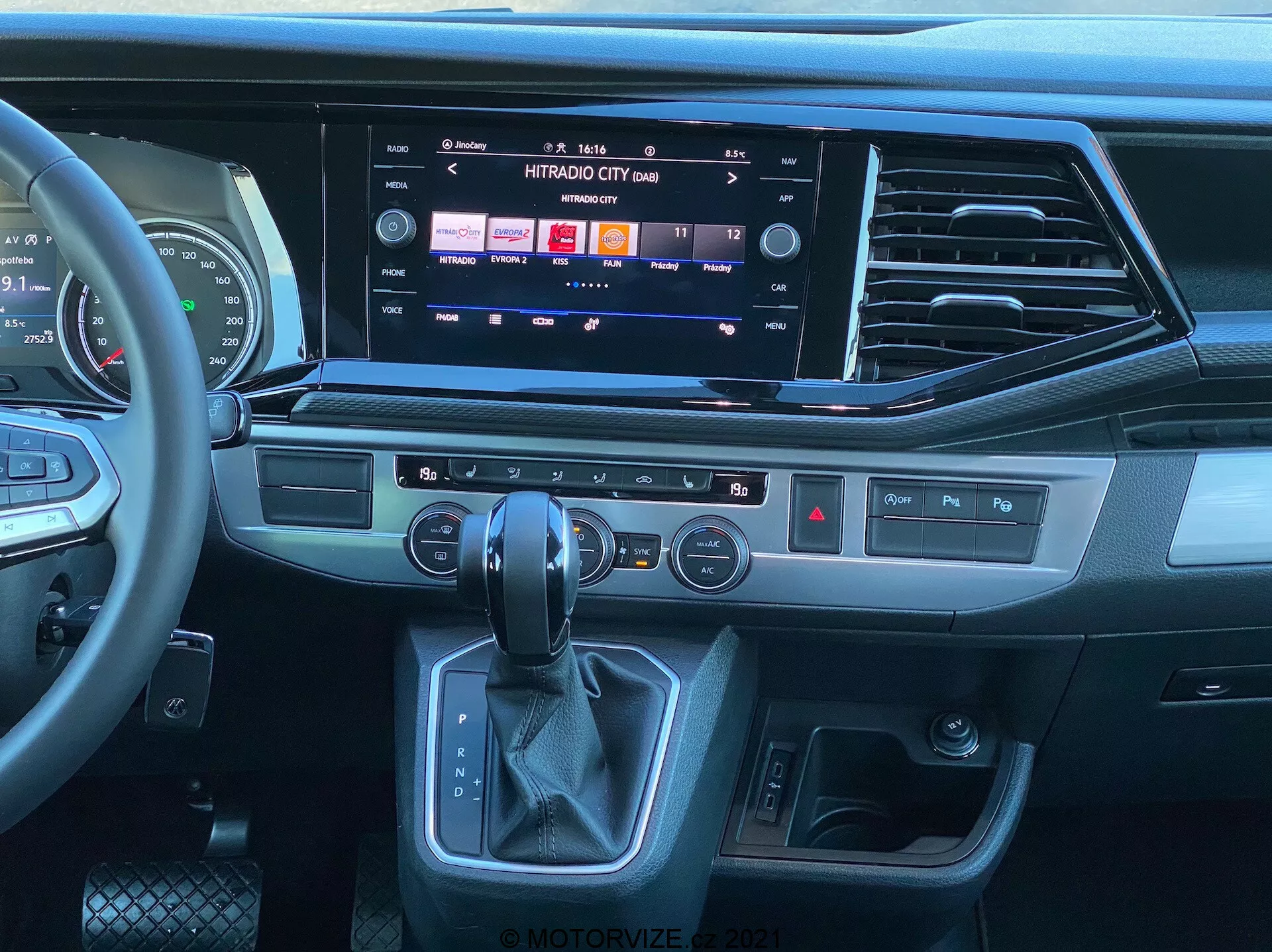 Vue de l'intérieur du VW Nutzfahrzeuge Transporter 2019 T6.1 Multivan depuis la perspective du siège passager, montrant le tableau de bord moderne et la console centrale. Volant multifonction en cuir avec commandes multimédias, grand système d'infodivertissement à écran tactile affichant des options multimédias et de connectivité, élégantes bouches d'aération, boutons tactiles pour fonctions rapides, panneau de commande de la climatisation bi-zone, levier de vitesse automatique ergonomique et bouton de démarrage/arrêt sans clé. Le design présente un mélange de plastiques sombres, de garnitures ressemblant à du métal brossé et de matériaux similaires au cuir, soulignant une ambiance contemporaine et premium.
