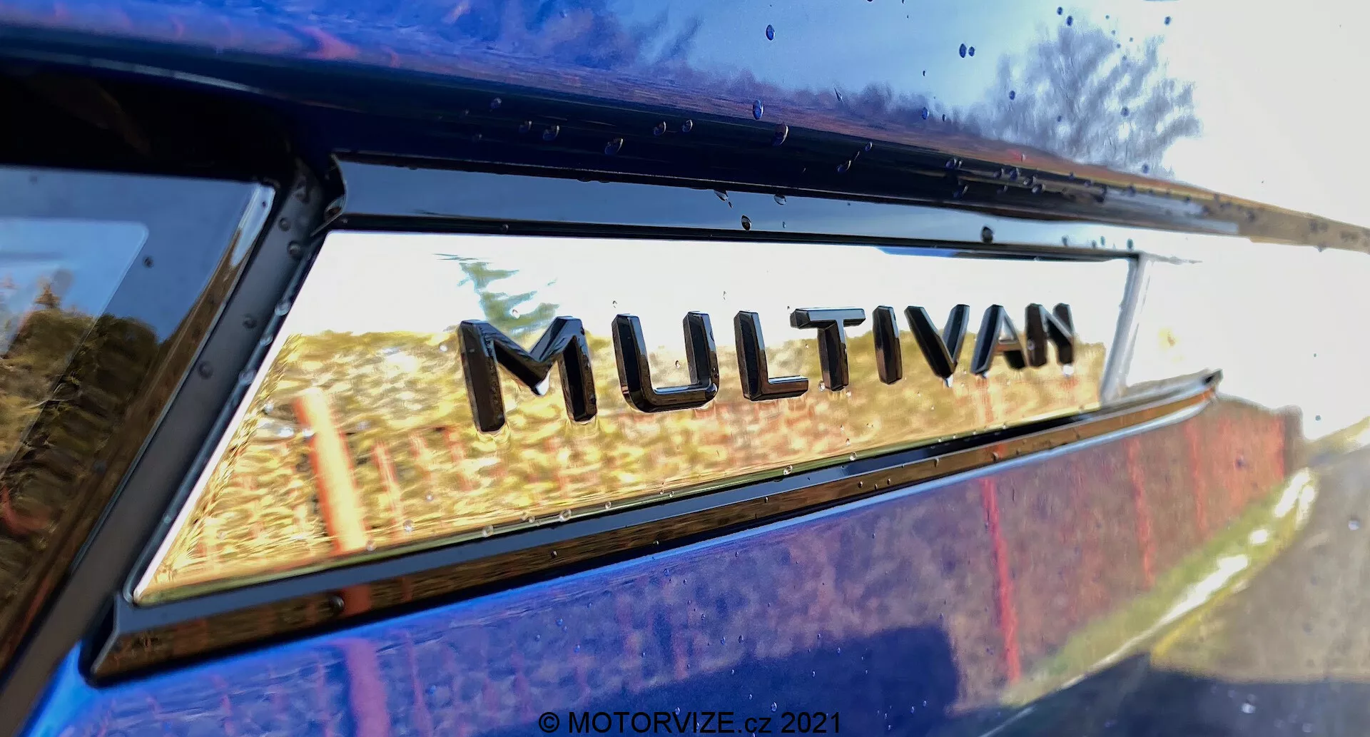 Vue rapprochée d'une partie du VW Nutzfahrzeuge Transporter 2019 (T6-1 Multivan) se concentrant sur le badge 'MULTIVAN'. L'image est prise d'un angle arrière bas, regardant légèrement vers le haut là où la vitre arrière rencontre le bord du toit. De légers reflets sur le verre de la fenêtre suggèrent un environnement extérieur. Cette image étroitement recadrée met en évidence l'emblème de désignation du modèle du fabricant.