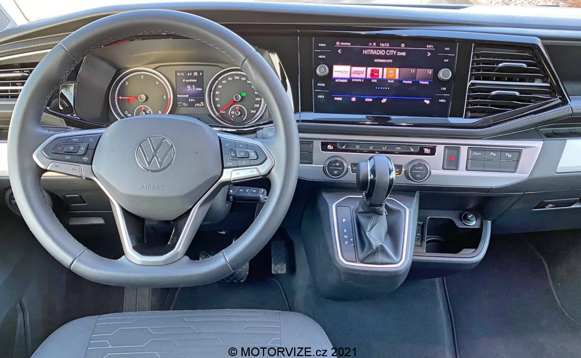 Vue intérieure du VW Nutzfahrzeuge Transporter 2019 T6.1 Multivan depuis la perspective du siège conducteur. Caractéristiques : volant arborant le logo Volkswagen avec boutons multifonctionnels, compteurs analogiques, écran numérique, console centrale avec système d'infodivertissement à écran tactile actif, climatisation bizone, levier de vitesse avec bouton de parking électronique et accents modernes en métal brossé.