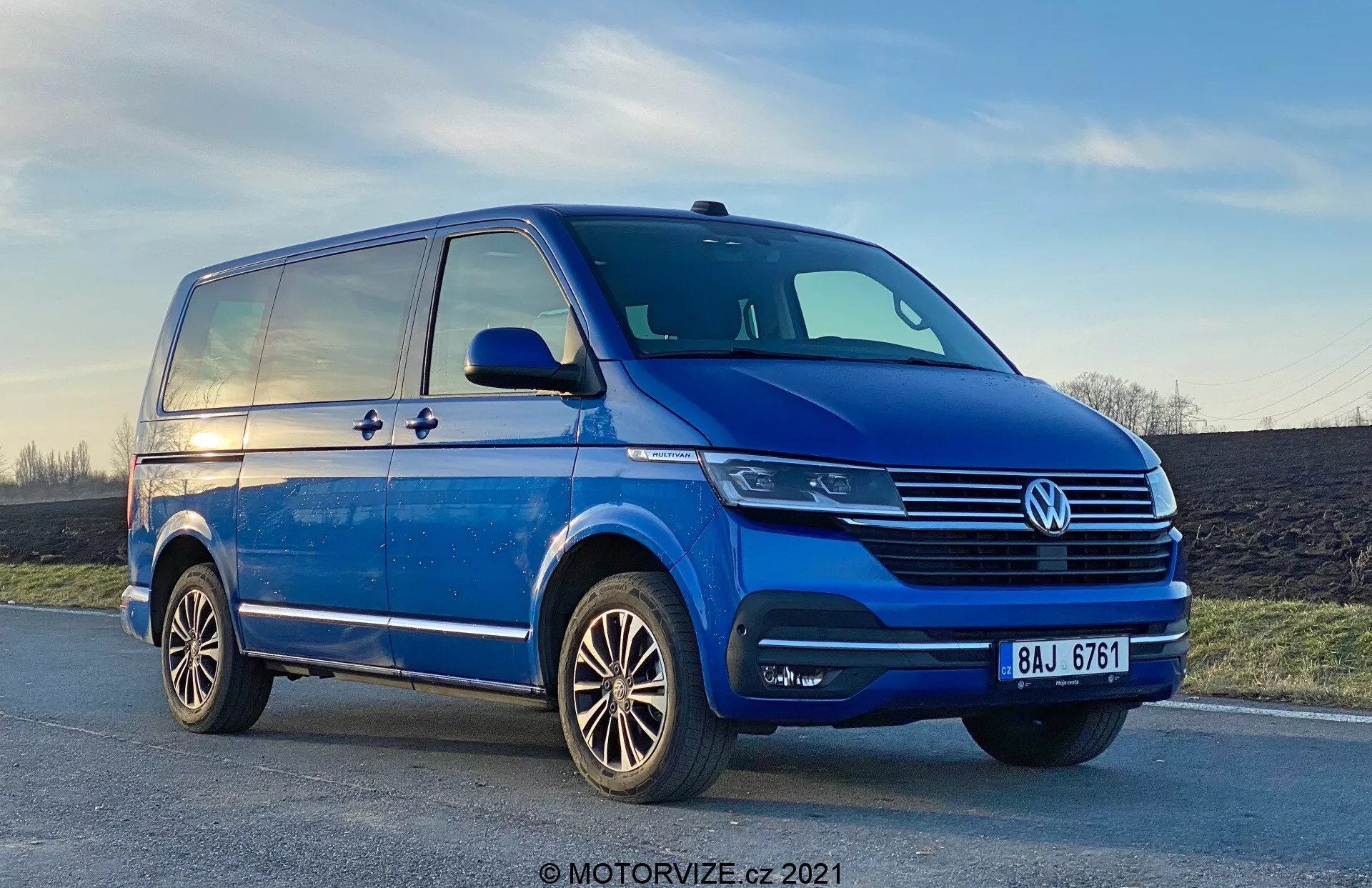 Vue en contre-plongée d'une Volkswagen T6-1 Multivan bleue de 2019, montrant le quadrant avant gauche d'une perspective trois-quarts. La camionnette est garée sur une route légèrement inclinée, avec des détails chromés proéminents sur la calandre, un emblème VW central et des unités de phares modernes qui pourraient inclure une technologie LED. Les feux antibrouillard sont intégrés dans le pare-chocs, qui dispose également de prises d'air. Le profil latéral révèle une porte coulissante, une ligne de caractère qui s'étend de l'aile avant à l'arrière et des rétroviseurs latéraux substantiels avec des indicateurs de virage. On peut également voir des roues en alliage avec un design multi-branches et des pneus bien entretenus. Le ciel en arrière-plan montre une touche de coucher de soleil.