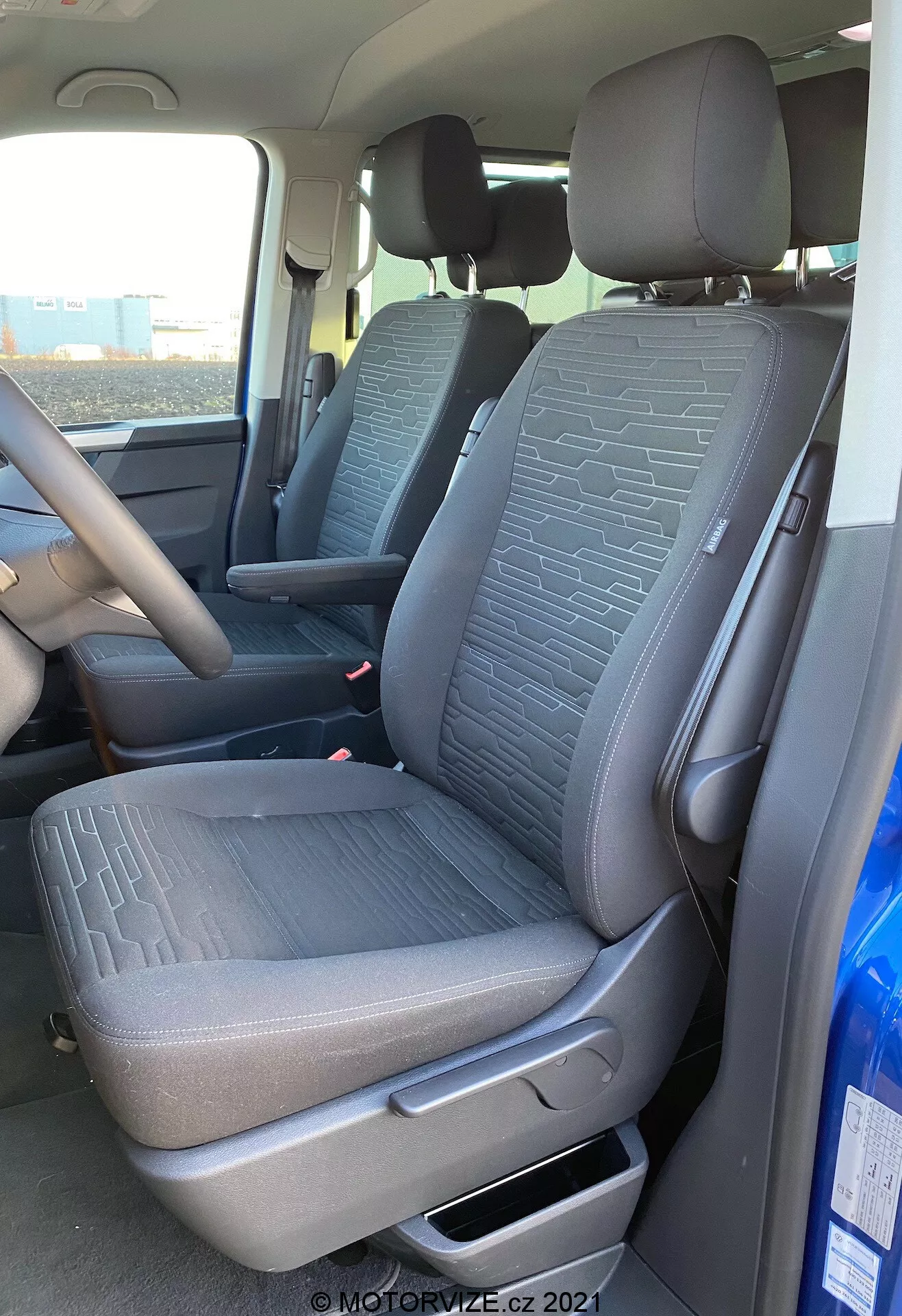 Intérieur du VW Nutzfahrzeuge Transporter 2019 T6.1 Multivan, montrant le siège passager avant et une partie du siège conducteur depuis une perspective extérieure du côté passager. La prise de vue, effectuée à un angle légèrement bas, met en évidence le tissu texturé avec des motifs géométriques sur les sièges, le levier de réglage manuel, les ceintures de sécurité et une partie de la console centrale et du volant. La lumière naturelle éclaire l'intérieur du véhicule.