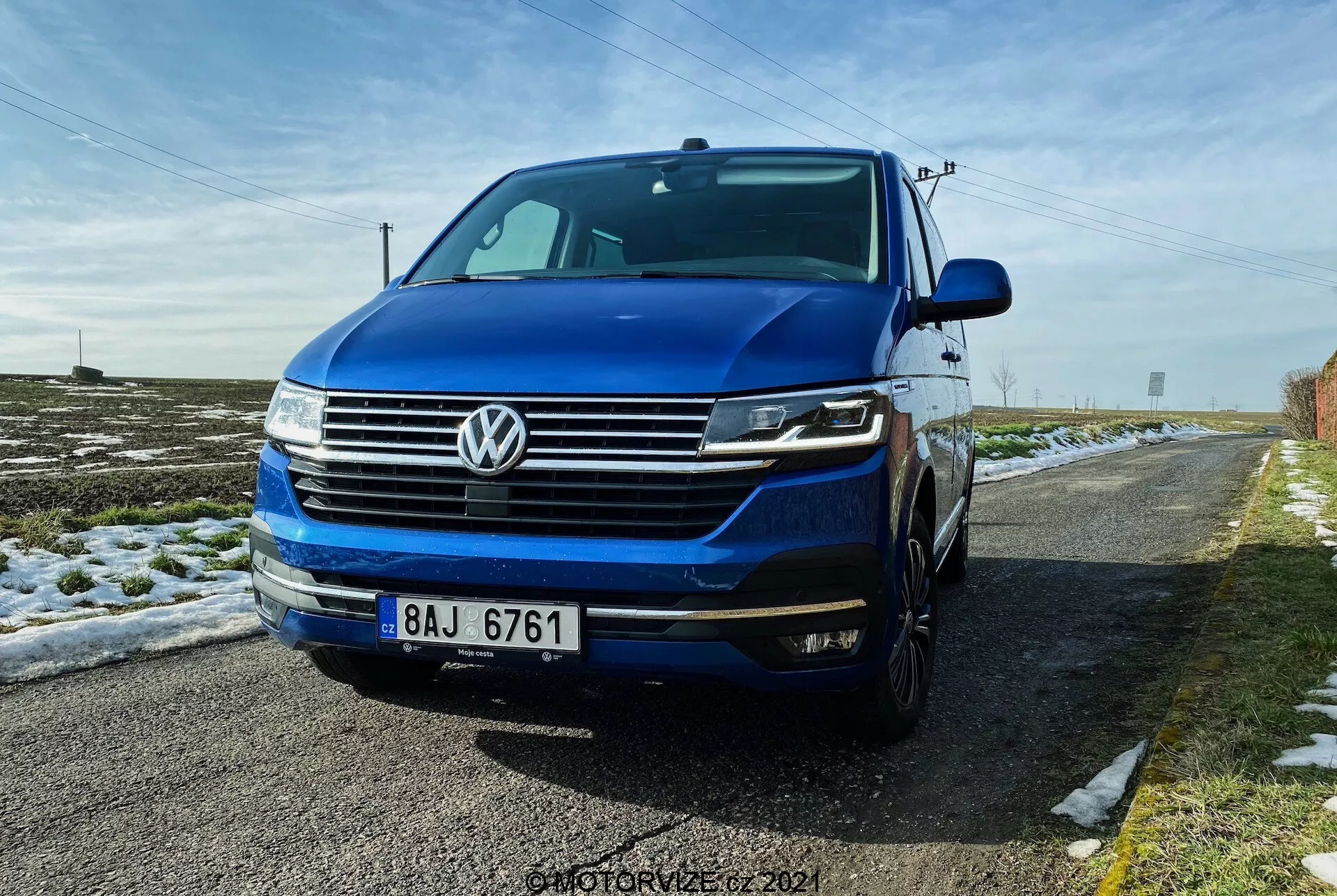 Vue avant trois-quarts basse du Volkswagen T6.1 Multivan 2019, avec la calandre avant à lignes horizontales, l'emblème distinctif VW centré sur la calandre, les phares probablement équipés de feux diurnes LED et de phares bi-xénon ou LED, et le pare-chocs avant avec des antibrouillards et des entrées d'air. L'image montre une partie du côté gauche, y compris la porte du conducteur et l'aile, avec le capot et le pare-brise visibles. La voiture est bleu foncé et garée sur une route rurale avec de la neige au sol et un ciel bleu clair au-dessus.