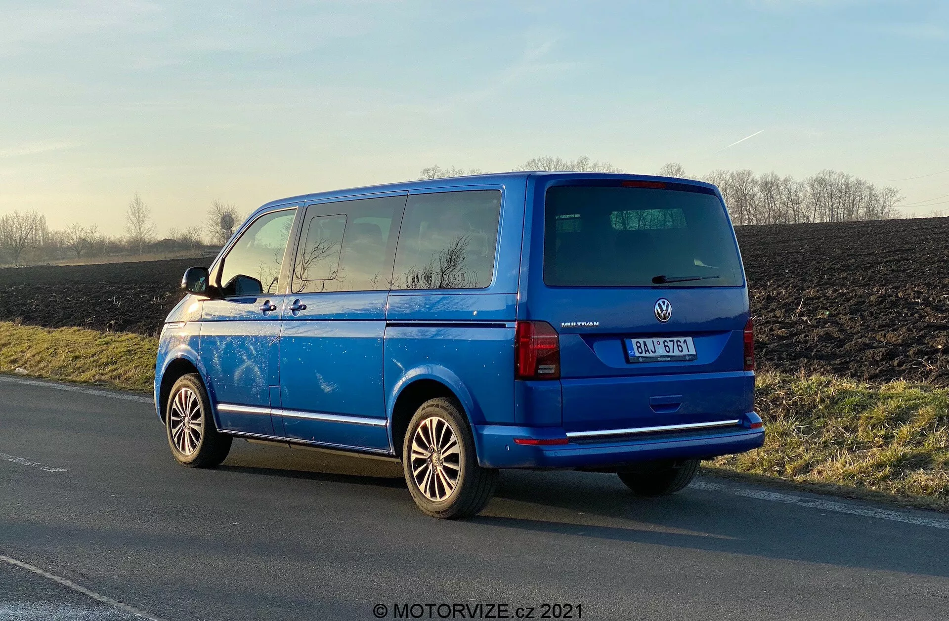 Volkswagen Transporter 2019 (T6-1 Multivan) bleu vibrant stationné sur le bas-côté, vue trois-quarts arrière montrant feux arrière, vitre arrière, porte arrière, badge du constructeur, détails du modèle du véhicule; roues en alliage proéminentes, rétroviseurs extérieurs, lignes du dégivreur de la vitre arrière, vitres teintées de la deuxième rangée, antenne sur le toit; stationné en plein jour avec des reflets soulignant la surface polie, arrière-plan rural.