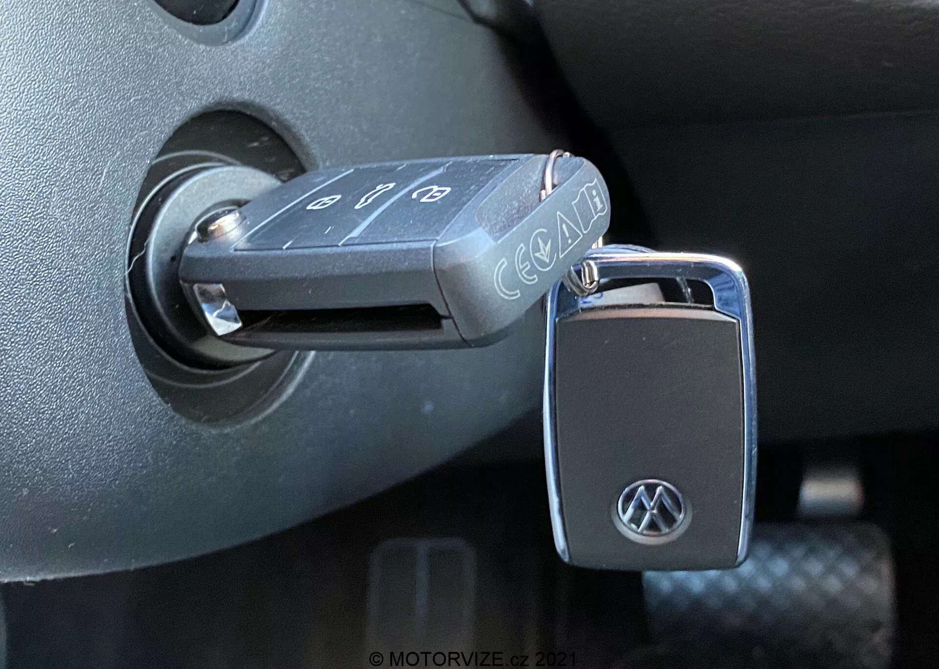 Gros plan sur une clé pliante Volkswagen insérée dans le contact d'un VW Nutzfahrzeuge T6-1 Multivan 2019. La clé, avec des boutons pour verrouiller, déverrouiller et alarme panique, arbore un logo VW sur le porte-clés. Le contacteur est situé dans l'environnement en plastique sombre de la colonne de direction, indiquant que l'allumage est en position 'éteint'. La photo, prise sous un angle latéral avec lumière naturelle, met en valeur les textures et les finitions détaillées de l'intérieur du véhicule.