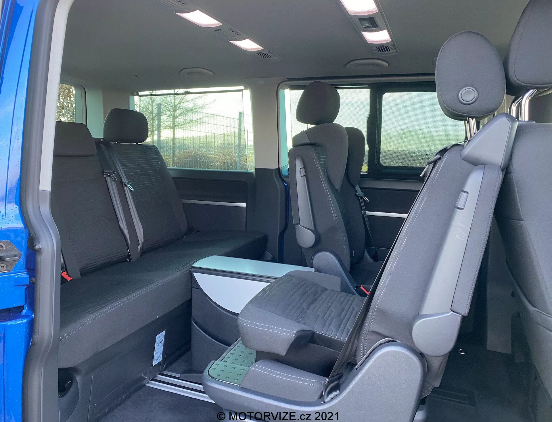 Intérieur du VW Transporter T6.1 Multivan 2019 montrant les sièges des deuxième et troisième rangées depuis la perspective du côté passager arrière, avec une configuration de sièges 2-3-2 et un revêtement en tissu foncé. Vue partielle de l'extérieur à travers la porte ouverte, appuie-tête sur tous les sièges et sièges de la troisième rangée surélevés pour une meilleure visibilité.