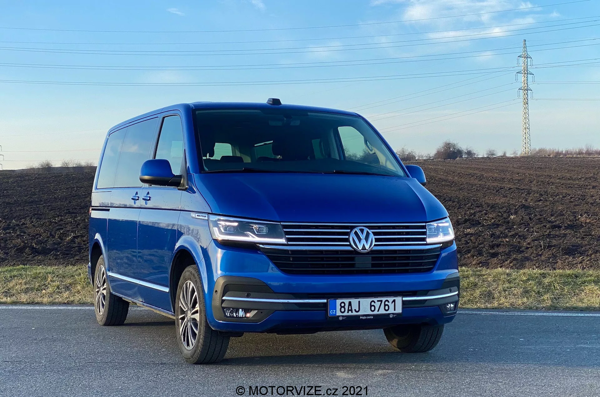 L'image présente une camionnette bleue Volkswagen VW-Nutzfahrzeuge Transporter 2019 (T6-1 Multivan) capturée sous un angle frontal bas, mettant en valeur le quart avant gauche avec une vue claire de sa façade avant et du profil côté conducteur. Le véhicule se trouve sur une route, mettant en évidence des éléments tels que l'emblème proéminent de Volkswagen, les phares élégants, les lignes sculptées du capot, les feux de brouillard intégrés au pare-chocs et les roues en alliage à multiples branches. La photographie, prise en pleine journée, souligne le style extérieur de la camionnette et les technologies d'assistance à la conduite, comme les capteurs de stationnement.