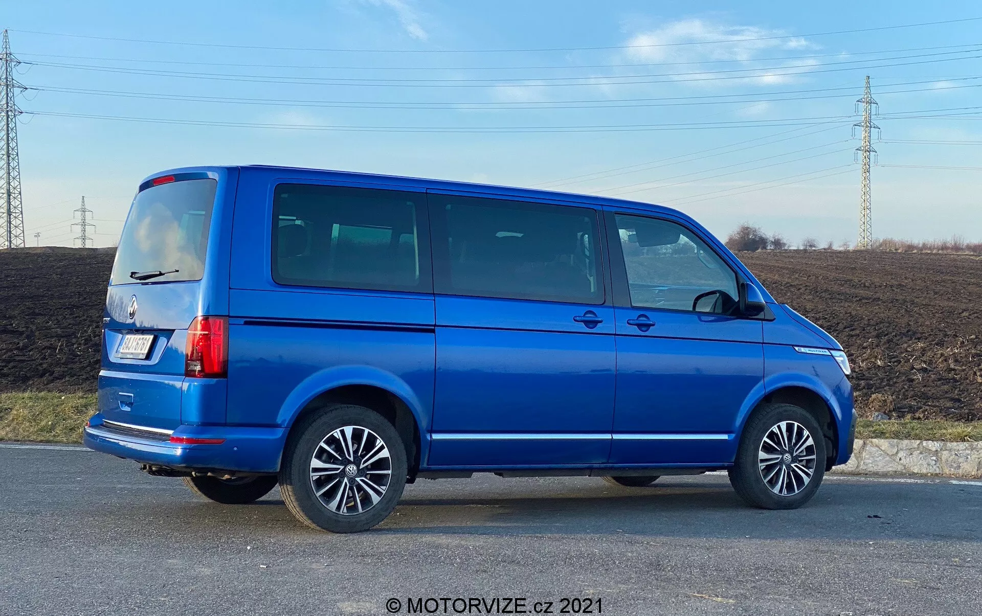 Vue de profil complet d'un VW Nutzfahrzeuge Transporter bleu de 2019 (T6.1 Multivan) avec des roues en alliage, des lignes de porte visibles, des vitres latérales teintées, une silhouette carrée, des feux arrière verticaux, une antenne sur le toit et une peinture métallique bleue brillante. L'image est prise sous un angle offrant une perspective à la fois sur le côté et l'arrière du véhicule, située dans une zone rurale avec un champ et des lignes électriques en arrière-plan, sous une lumière de jour.