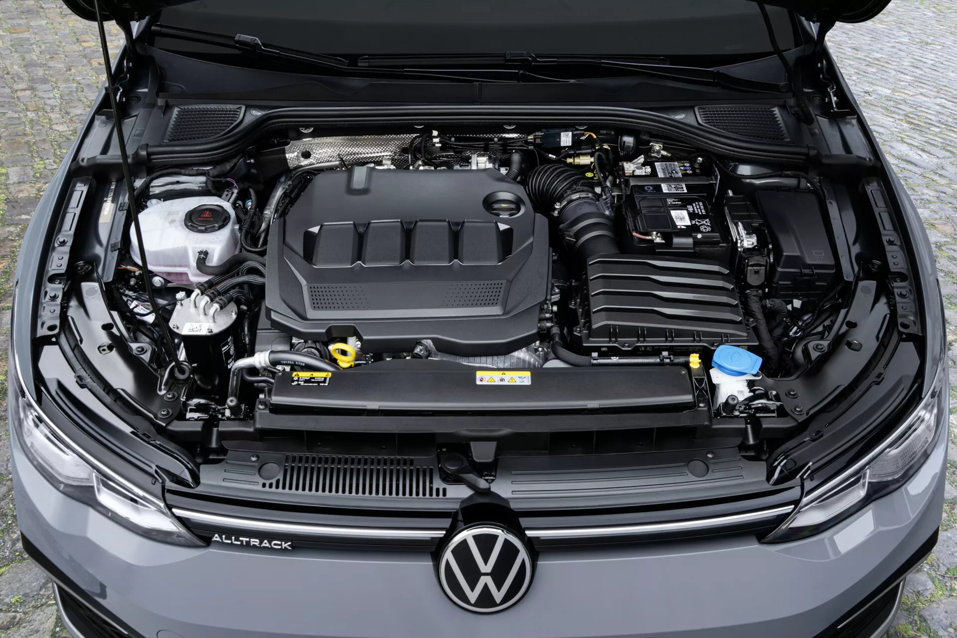 Vue en plongée sur le compartiment moteur d'une Volkswagen Golf Alltrack 2017 montrant le couvercle du moteur avec logo, réservoirs de fluides et batterie de voiture