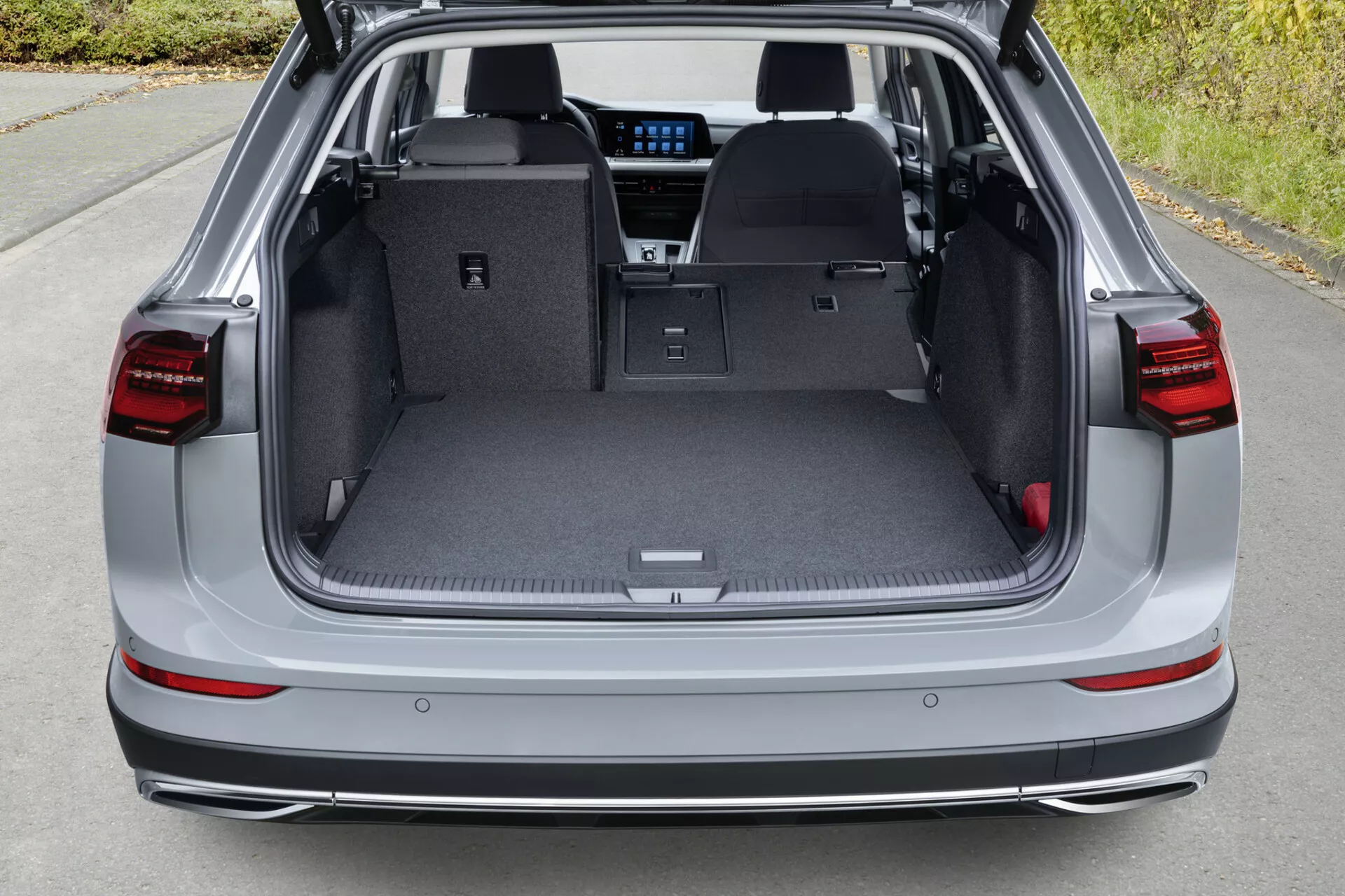 Vue en plongée de l'espace de chargement arrière d'une VW Golf Alltrack de 2017 avec le hayon ouvert, montrant le plancher de chargement plat avec les sièges arrière rabattus, les feux arrière LED, les capteurs de stationnement sur le pare-chocs arrière et des aperçus de la technologie d'infodivertissement moderne à travers l'arrière.