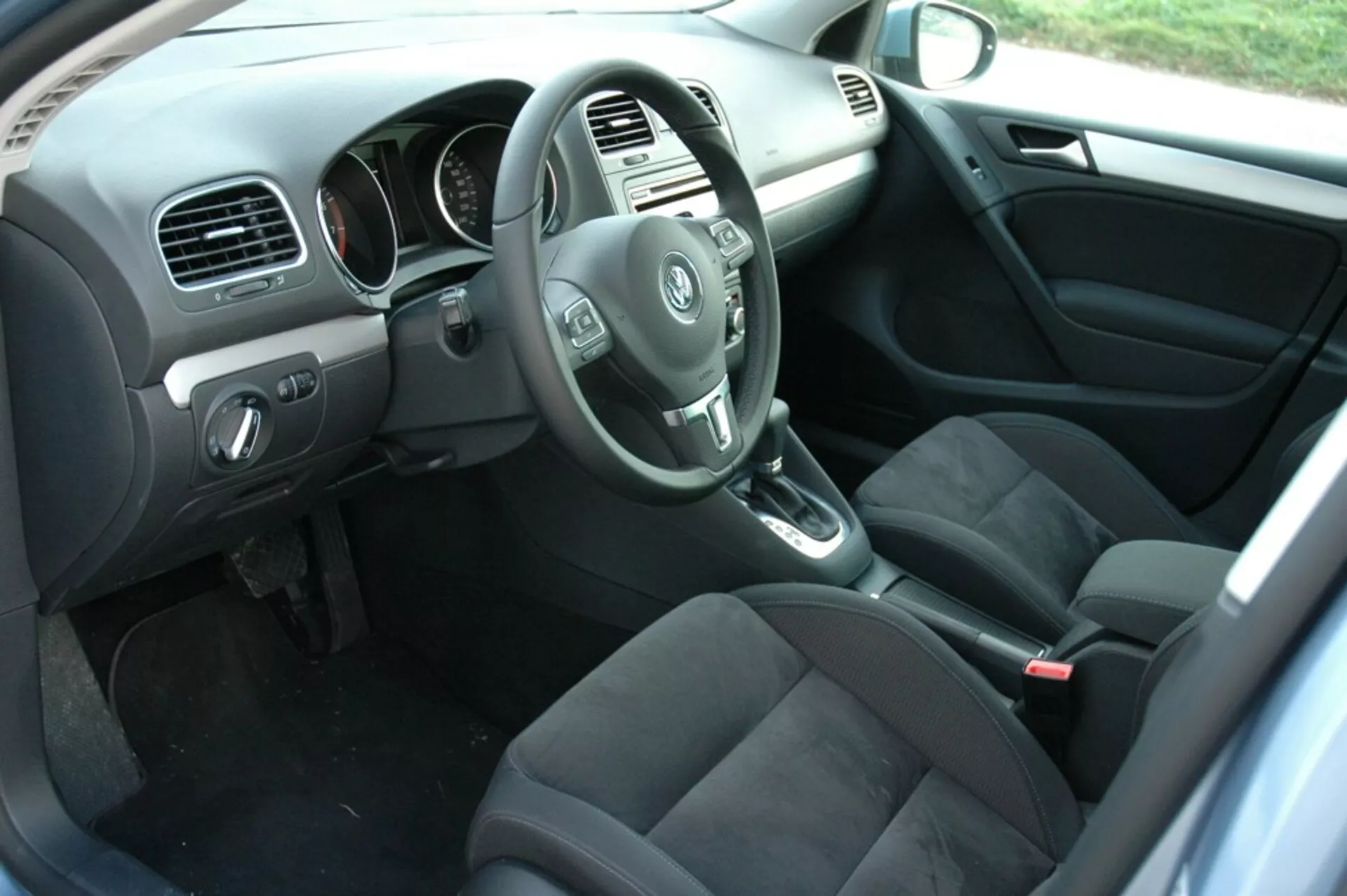 Intérieur de la VW Golf 2008 montrant le côté du conducteur depuis la perspective du passager arrière avec le volant, le tableau de bord, la console centrale, le levier de vitesse automatique, les sièges en tissu foncé, les commandes sur la porte du conducteur, le sol moquetté avec tapis et l'éclairage naturel