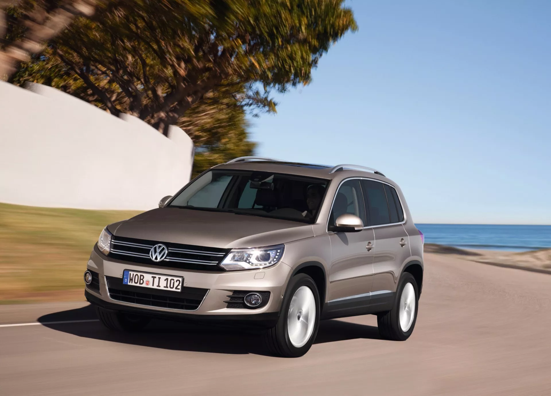SUV de taille moyenne Volkswagen Tiguan de 2007 roulant sur une route côtière, mettant en évidence sa grille avant, ses phares, son profil latéral et le design de ses jantes sous des conditions ensoleillées, indiquant un mouvement dynamique et une technologie de détection automatique de la lumière