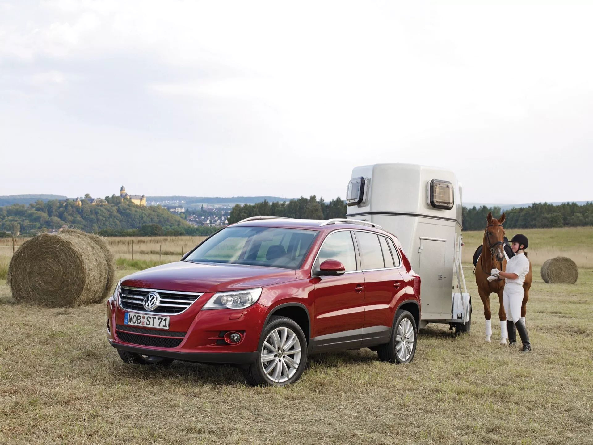 SUV rouge VW Tiguan de 2007 équipé de phares à faisceau projecteur et de roues en alliage, remorquant une remorque à chevaux dans un champ herbeux avec un château en arrière-plan à l'aube, mettant en lumière son système AWD et ses barres de toit pratiques.