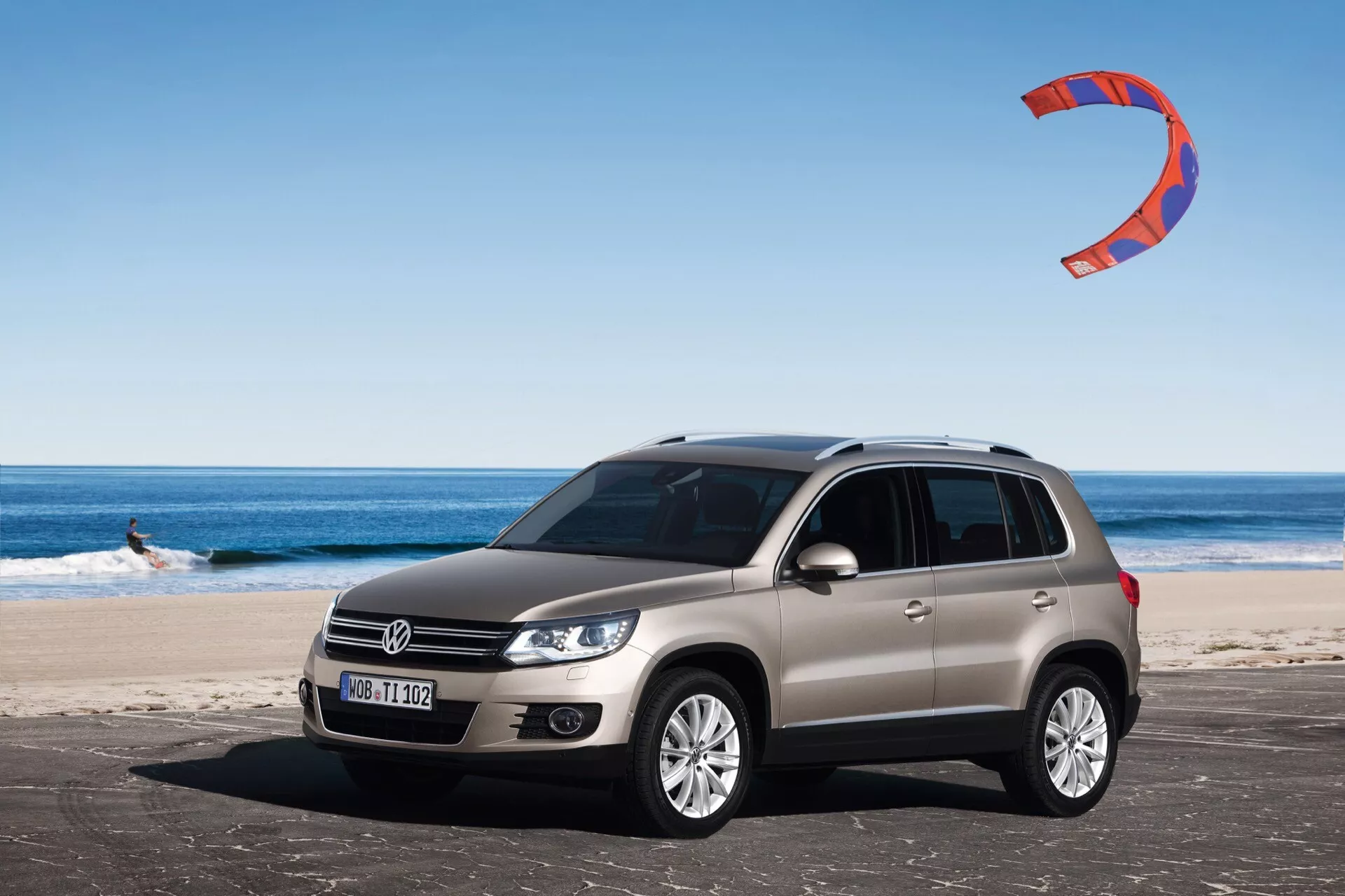 Vue avant trois quarts d'un SUV Volkswagen Tiguan de 2007 en argent, garé sur une plage de sable avec logo visible sur la calandre, phares halogènes, jantes en alliage à rayons multiples, et un kitesurfeur en arrière-plan sous un ciel dégagé