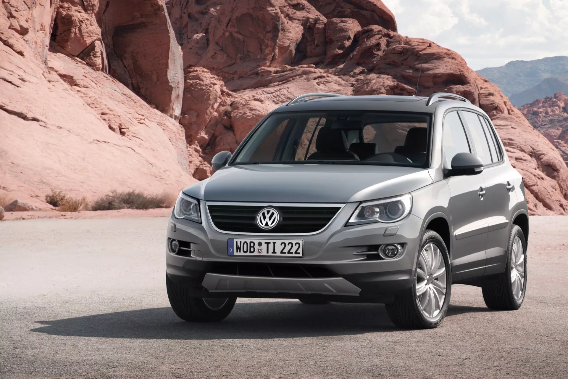 SUV VW Tiguan gris métallique de 2007 garé sur un terrain sablonneux avec des formations rocheuses en arrière-plan, vue du quart avant gauche montrant la grille, les phares, le pare-chocs avant avec feux de brouillard et capteurs, capot sculpté et roues en alliage