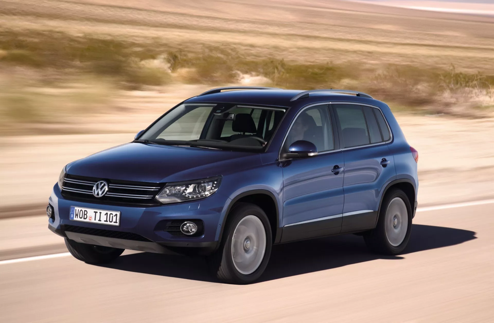 Volkswagen Tiguan 2007 en mouvement, capturée d'un angle avant trois-quarts montrant le côté conducteur, avec des caractéristiques telles que des roues en alliage, des barres de toit, des phares allumés, sur un fond de terrain plat et aride
