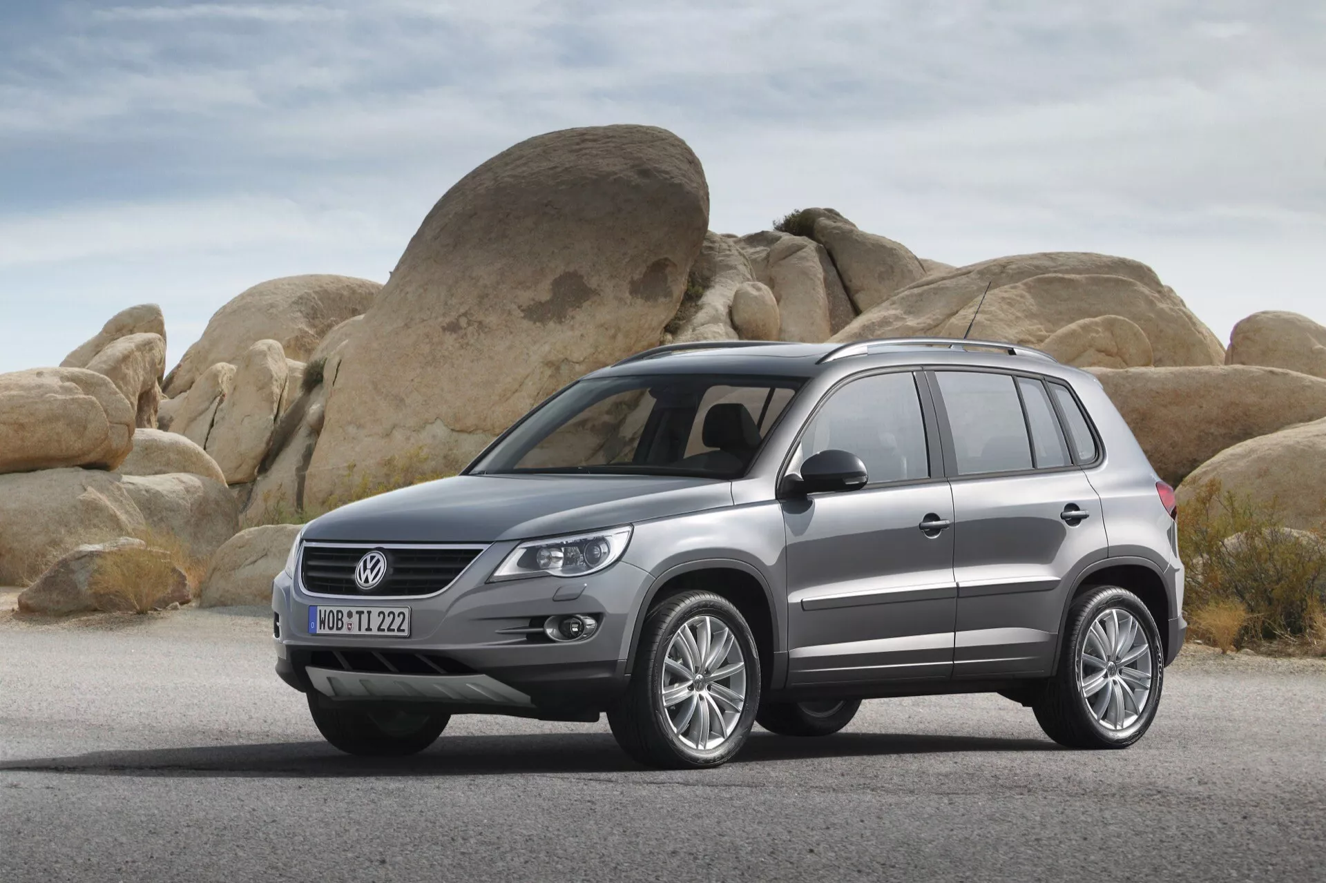 SUV Volkswagen Tiguan 2007 garé sur une surface sèche et plate avec de gros rochers arrondis en arrière-plan, mettant en avant le profil avant et latéral du véhicule incluant la grille, les phares, le pare-chocs avant, le capot, les roues, les panneaux latéraux, les portes et les vitres latérales avec des roues en alliage, des barres de toit et une finition peinture métallique.