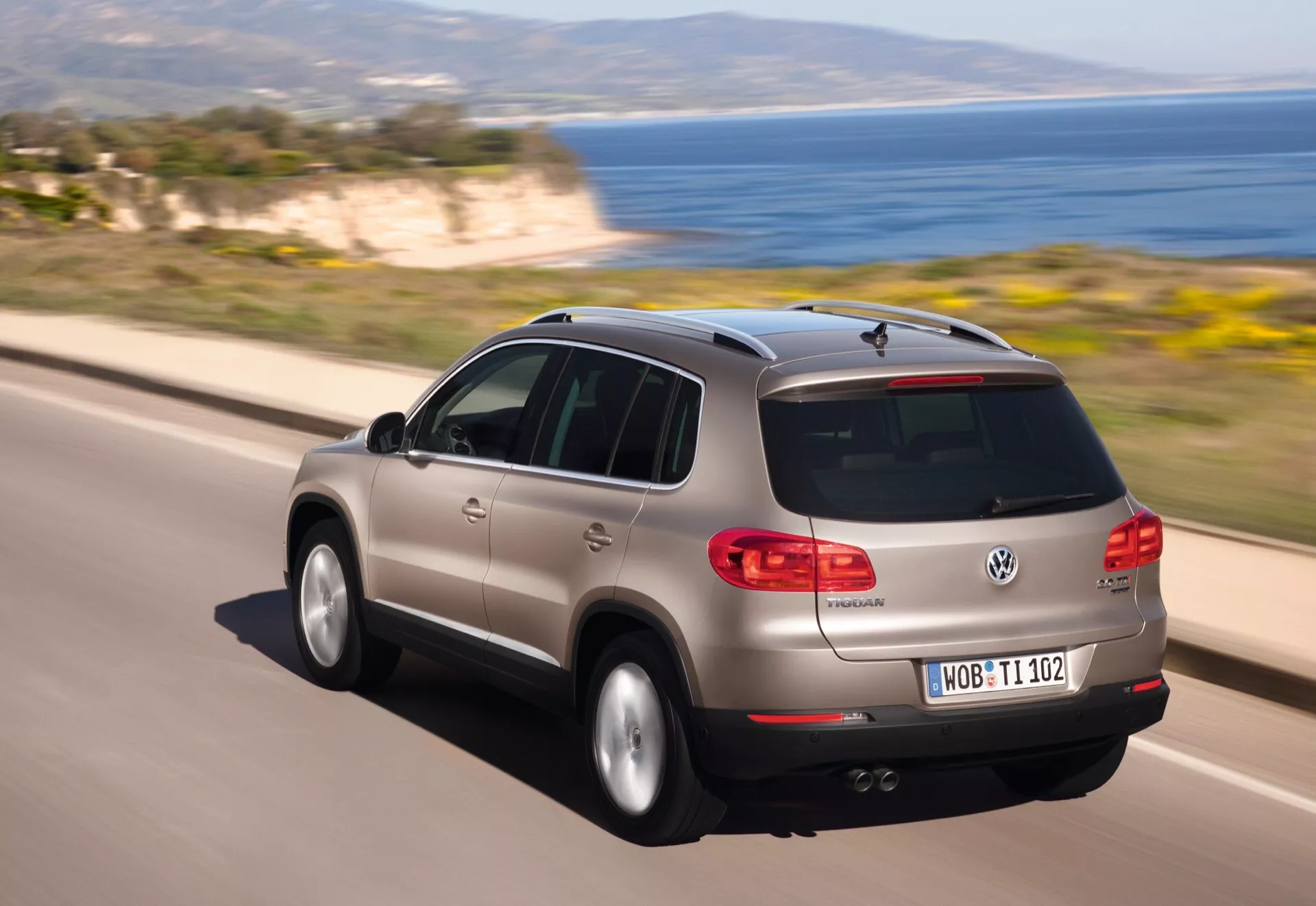 Vue arrière dynamique aux trois quarts d'une Volkswagen Tiguan argent en mouvement sur une route côtière, mettant en évidence son panneau arrière, le design du hayon, les feux arrière LED et les caractéristiques aérodynamiques telles que la vitre arrière inclinée, le spoiler au-dessus de la vitre arrière et l'antenne aileron de requin.